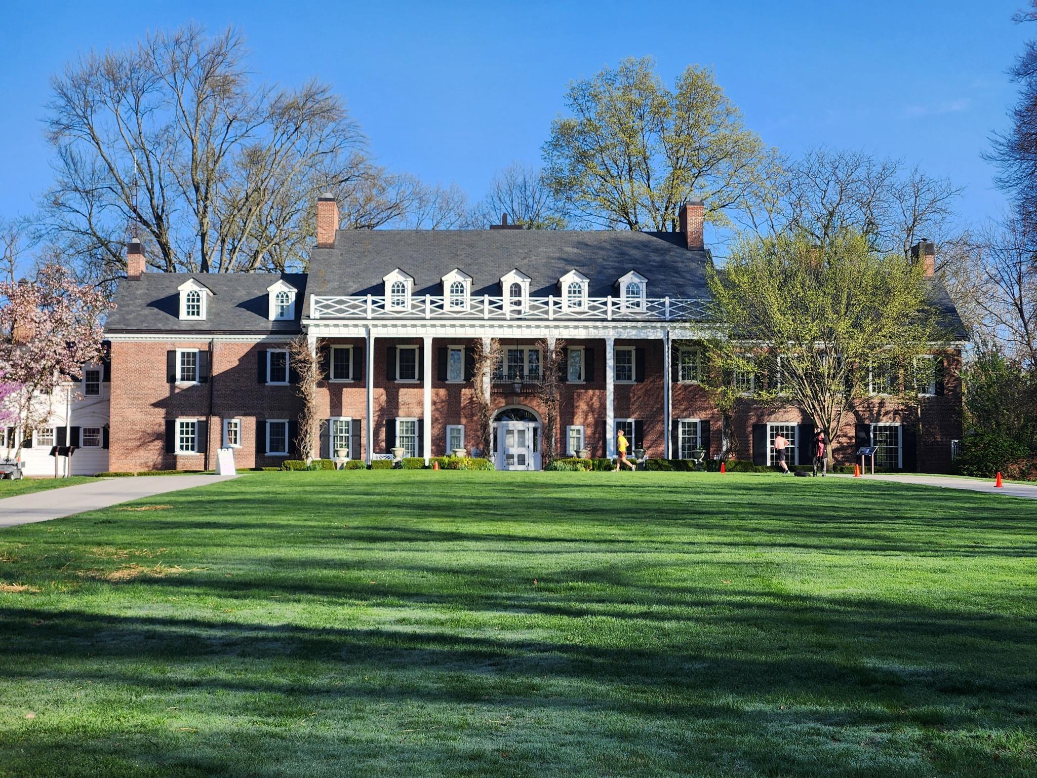 A grand brick mansion with white columns stands on a vast green lawn.
