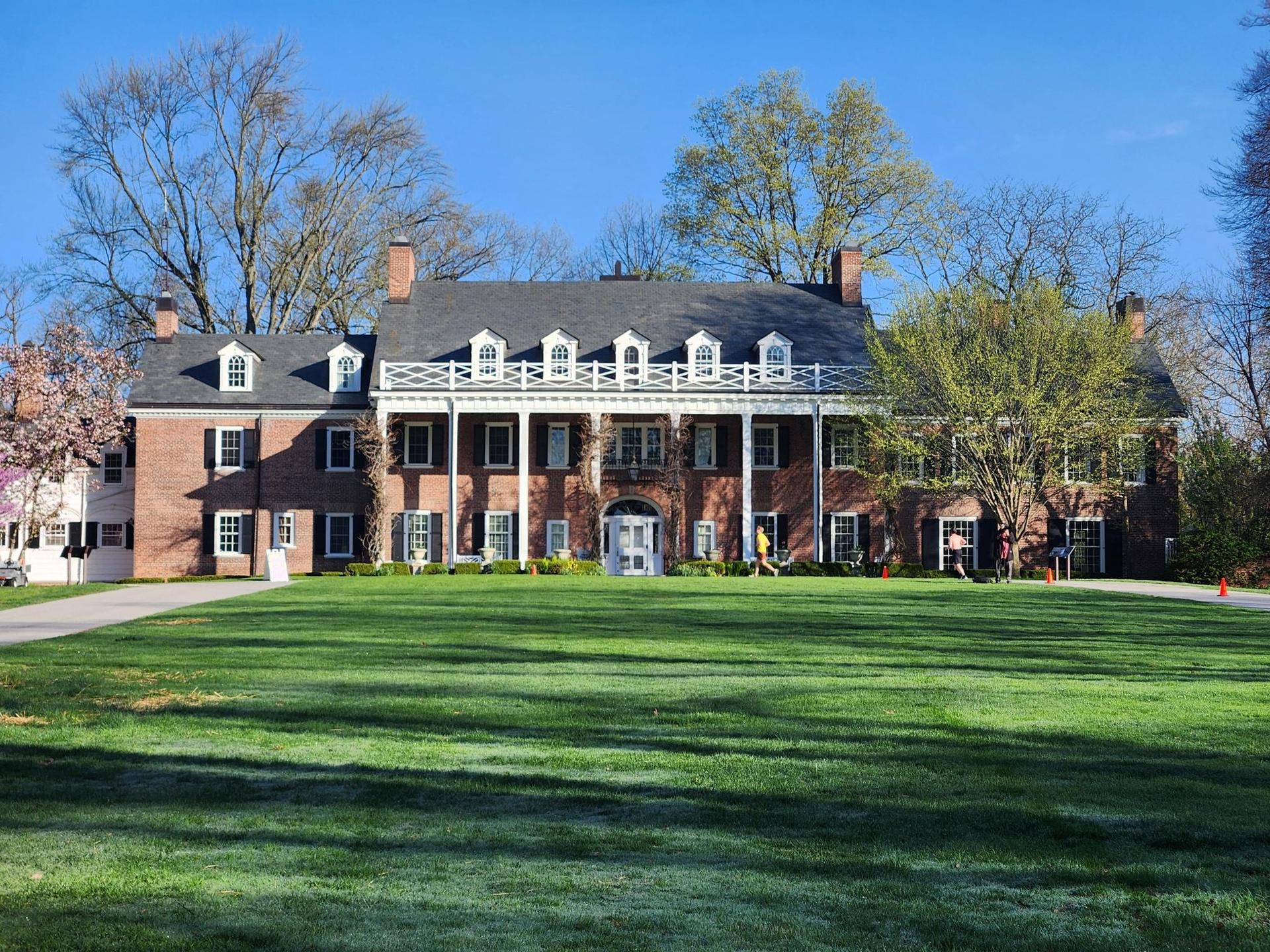 A grand brick mansion with white columns stands on a vast green lawn.