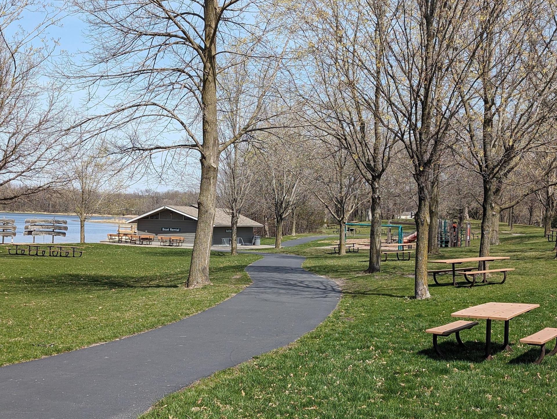 Park with picnic tables, bare trees, a building, and a lake.