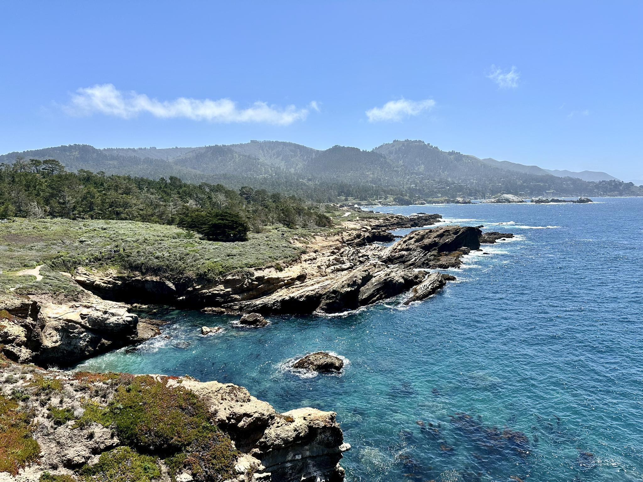 Clear turquoise ocean water meets a rocky, green coastline with distant mountains.