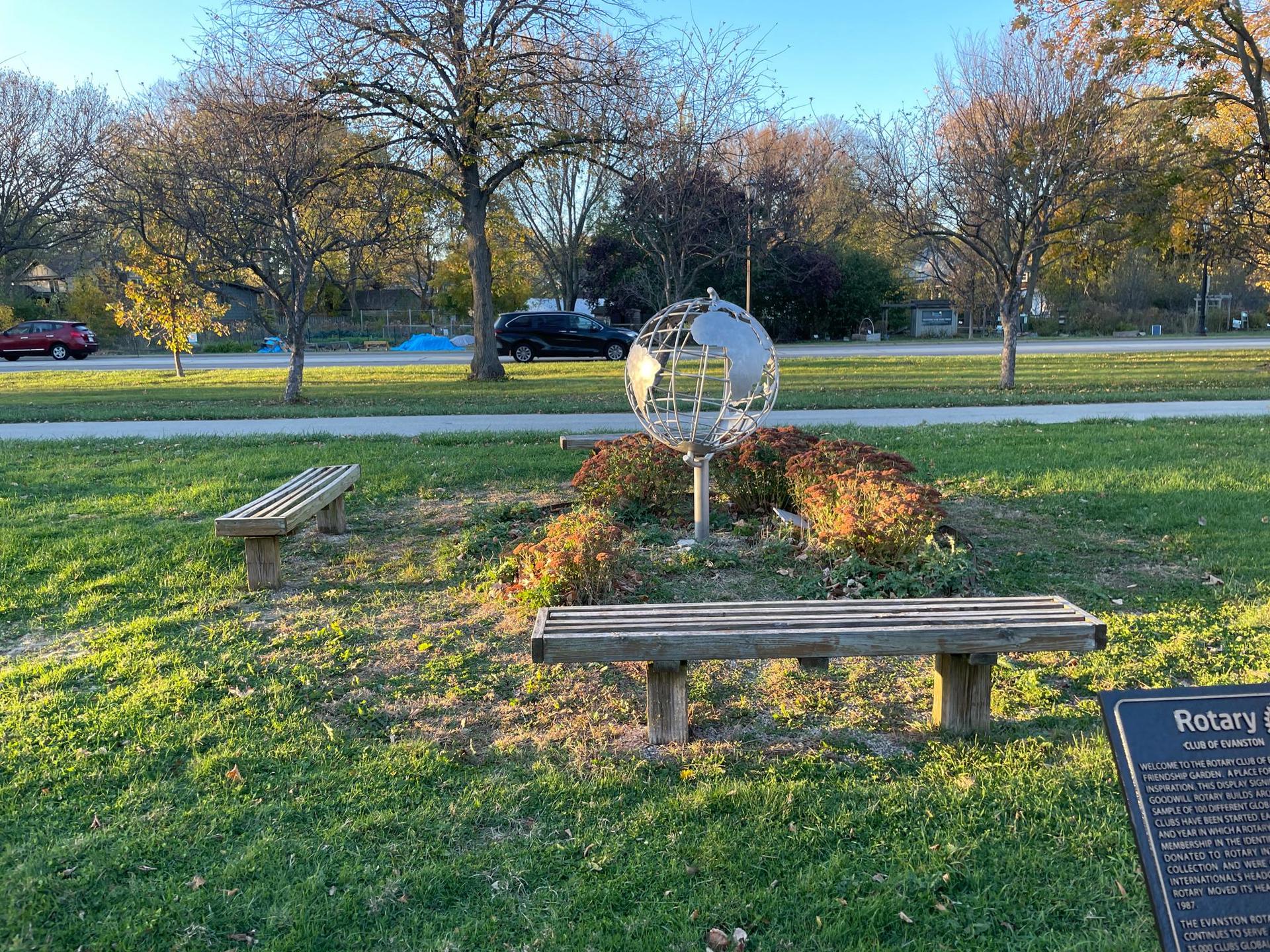 Two park benches flank a metal globe sculpture on a sunny, grassy lawn.