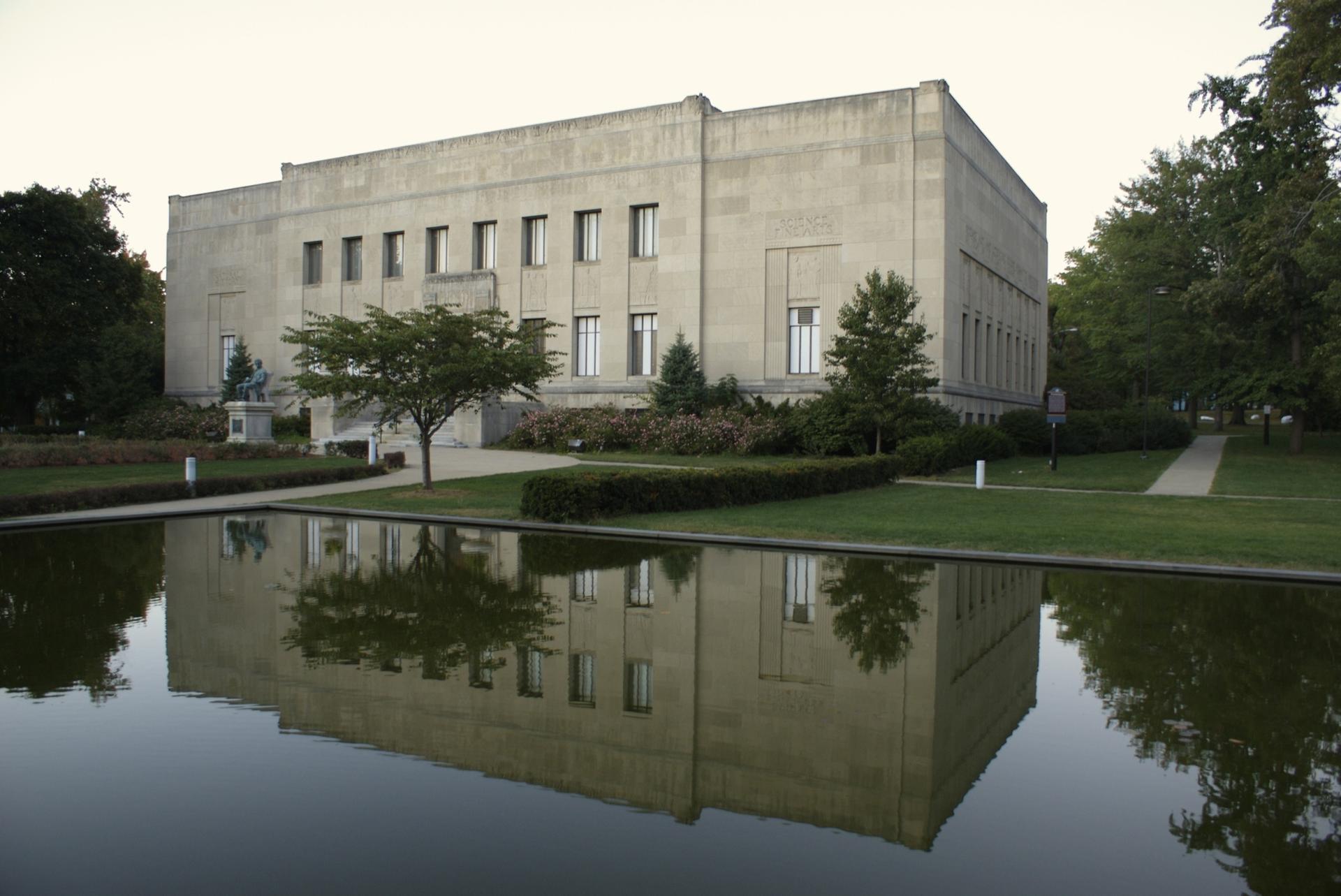 Large concrete building reflected in a calm pond, surrounded by green trees and grass.