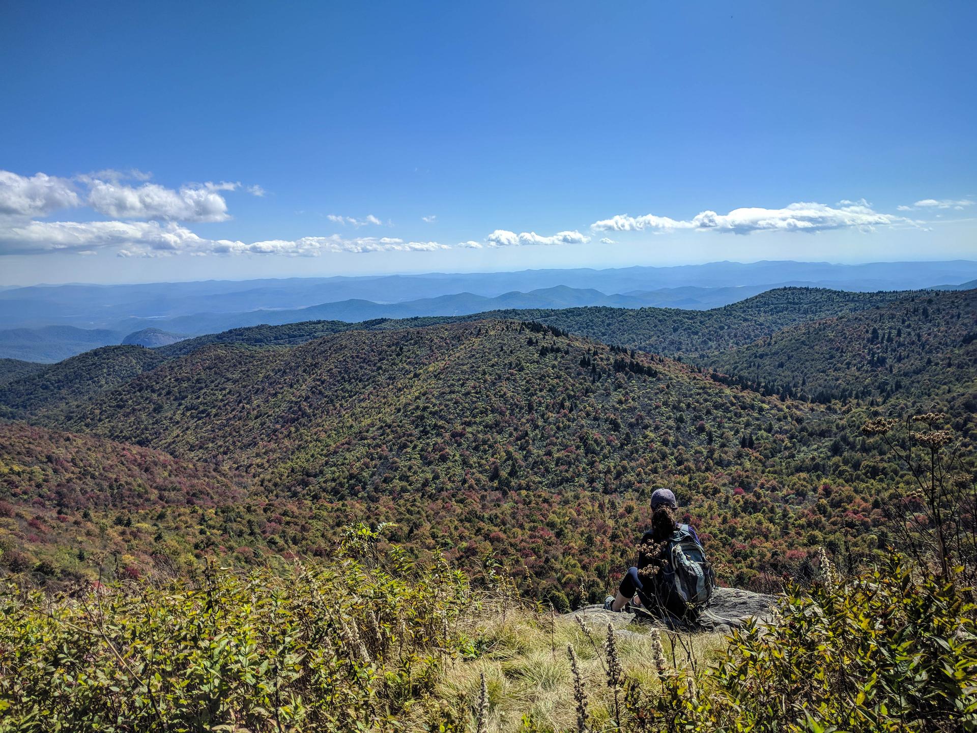 Person sitting on a rock, admiring vast mountain views under a blue sky.