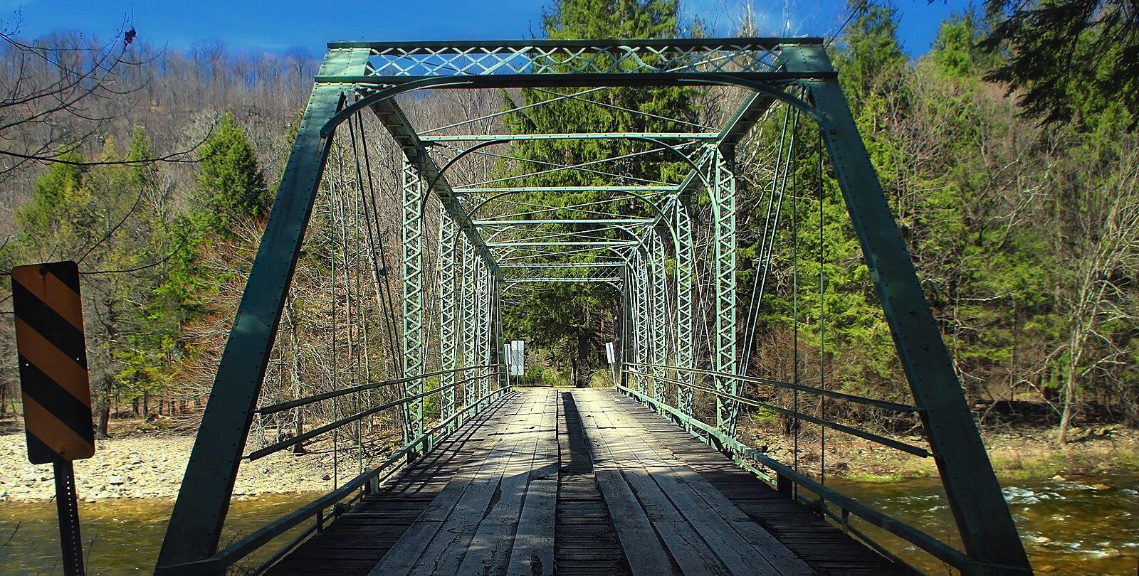 A green metal truss bridge with a wooden deck spans a river.