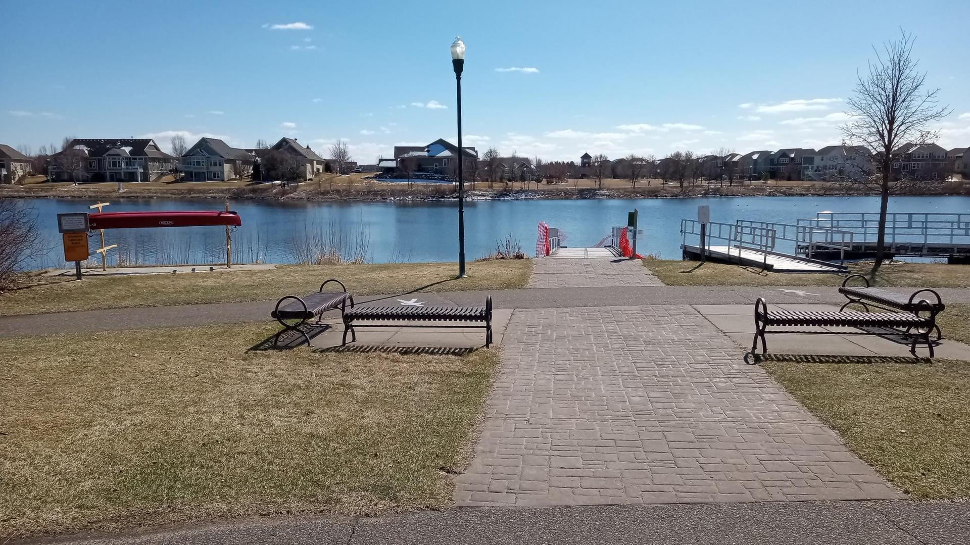 Paved path leading to a lake with a dock, benches, and distant houses.