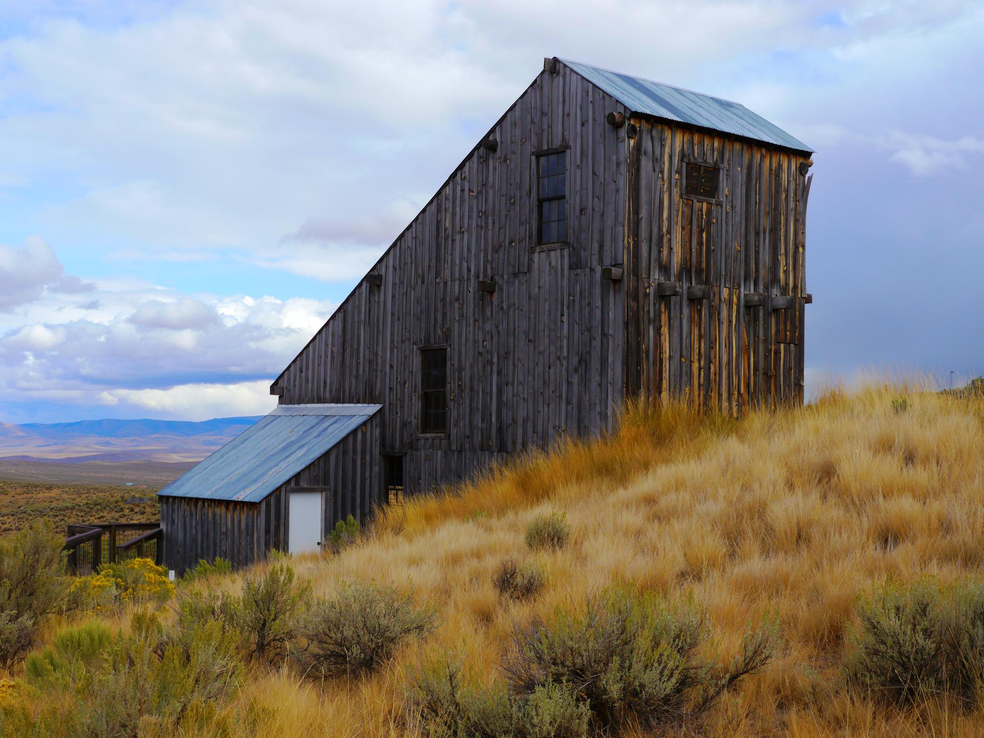 Weathered wooden building on a golden grassy hill with distant mountains under a cloudy sky.