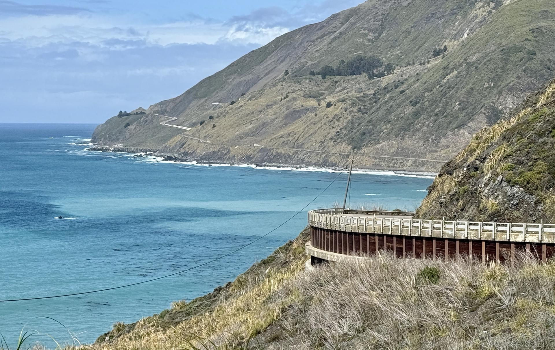 Curved coastal road with a railing overlooking the vast blue ocean and mountains.