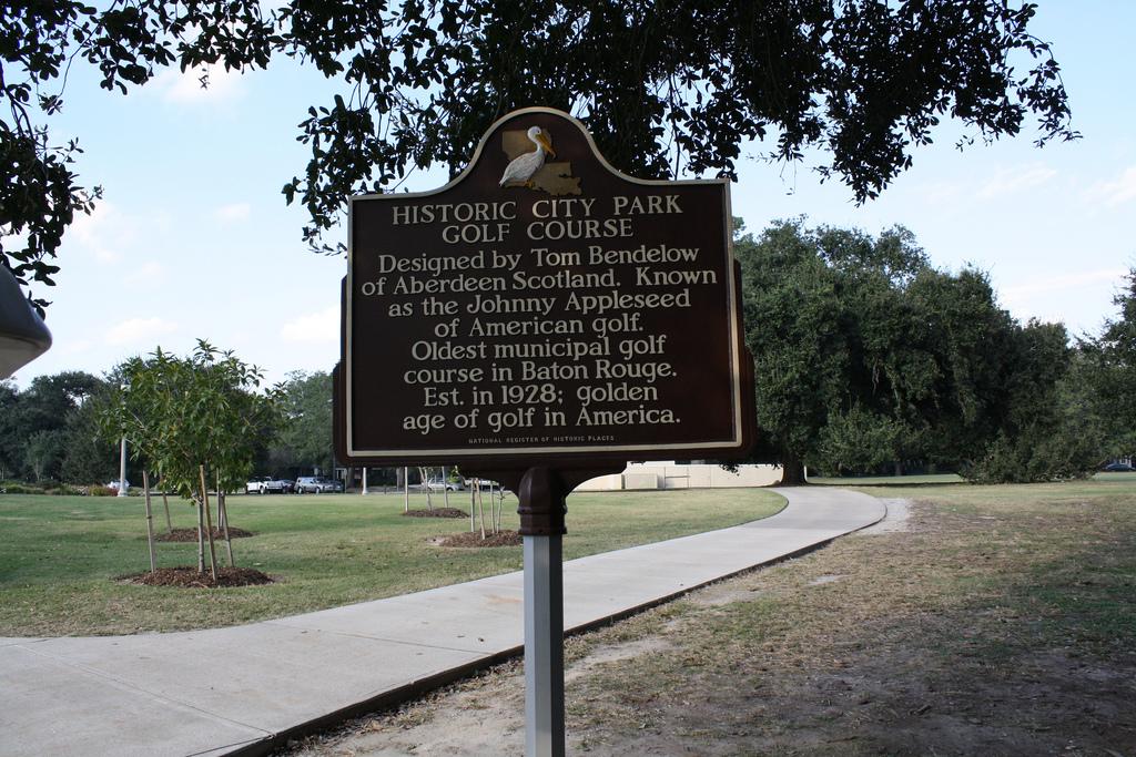 A brown historical marker stands beside a paved path in a sunny, grassy park.