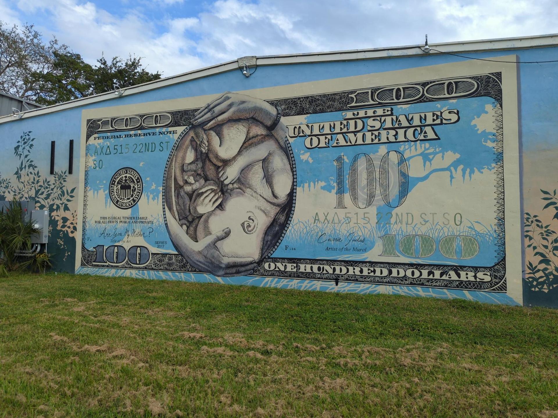 Large mural of a hundred-dollar bill with a baby on a building wall.