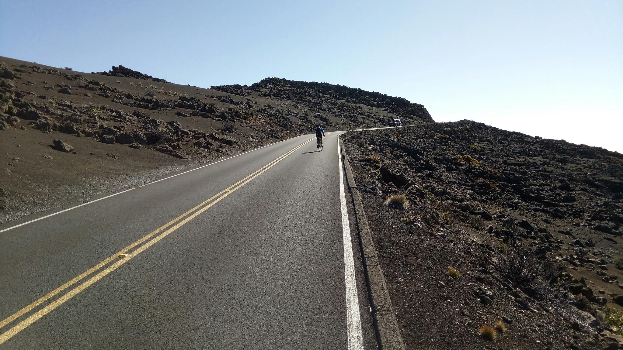 A lone cyclist rides uphill on a winding road through a barren, rocky landscape. - North Shore Greenway