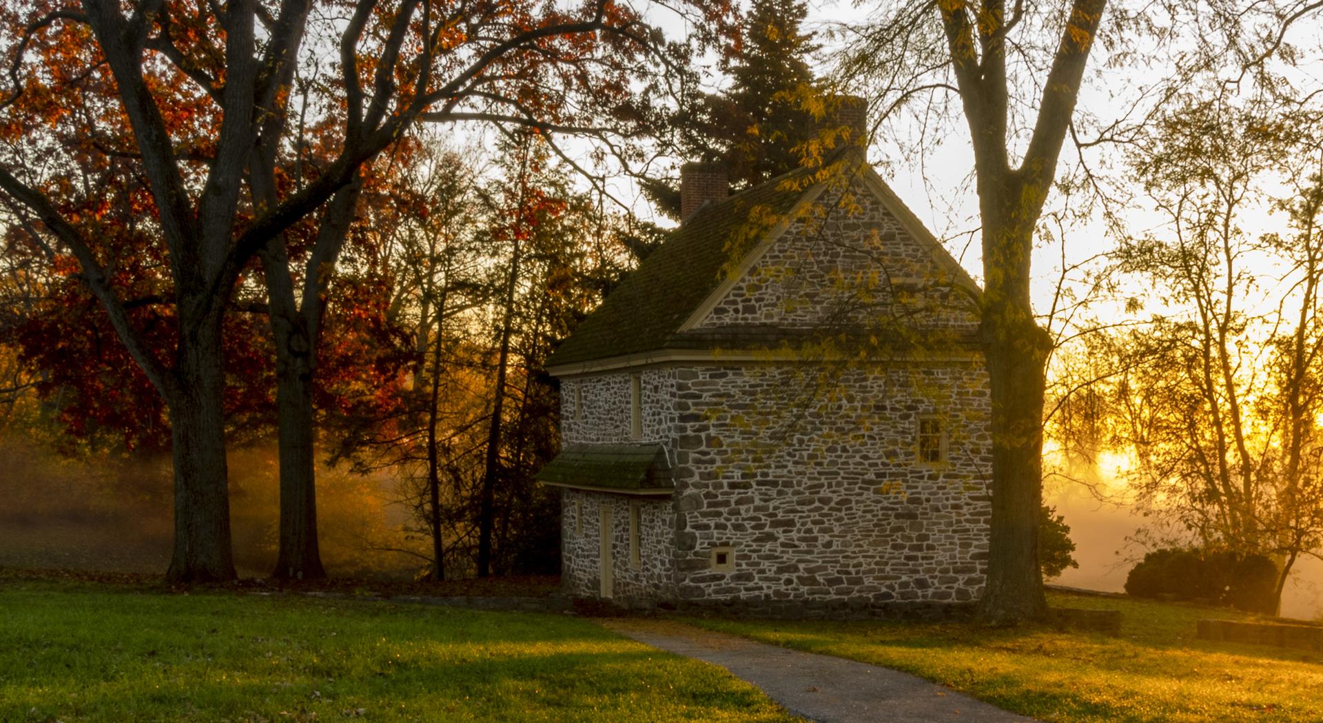 Stone house with a path, surrounded by trees with autumn leaves at sunset.