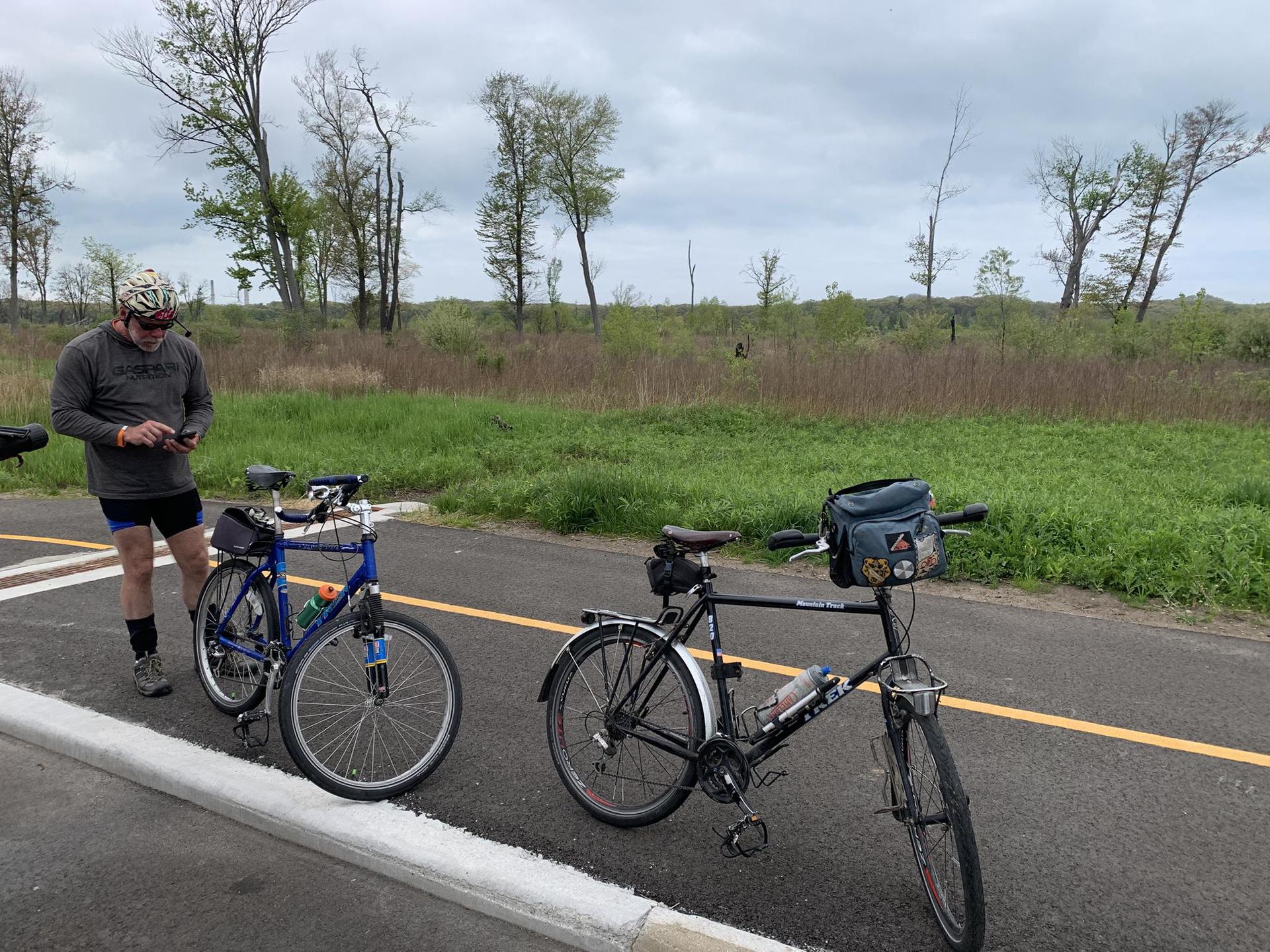 Cyclist checking phone beside two bikes on a paved path, overcast day.