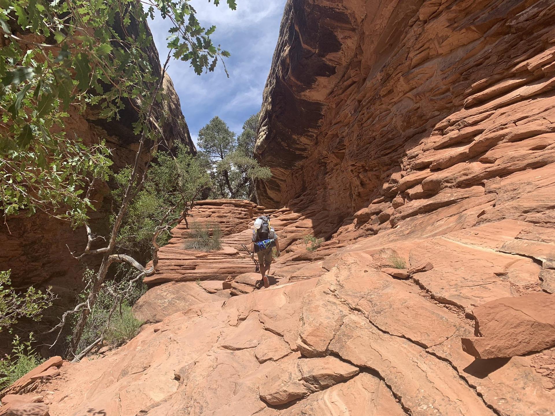 Hiker with backpack ascending a narrow, rocky red canyon path.
