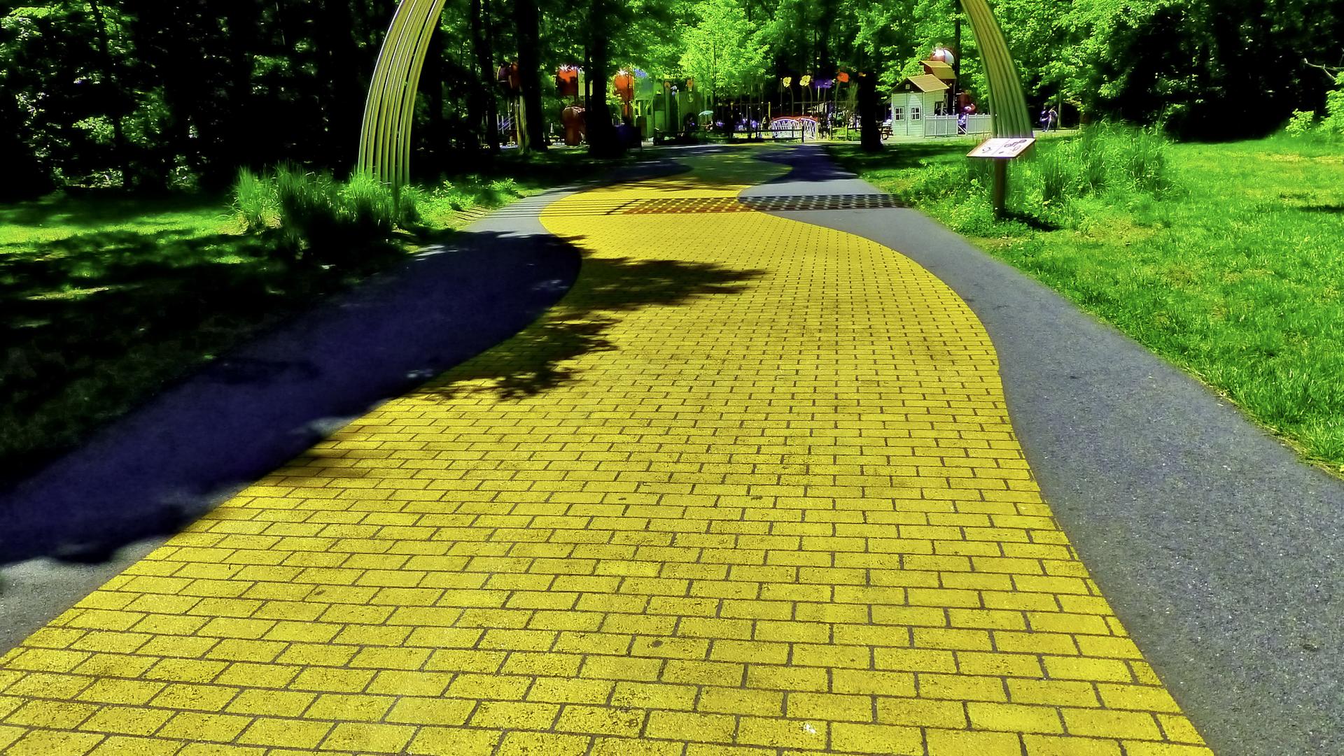 Bright yellow brick path curves through a sunny green park with trees.