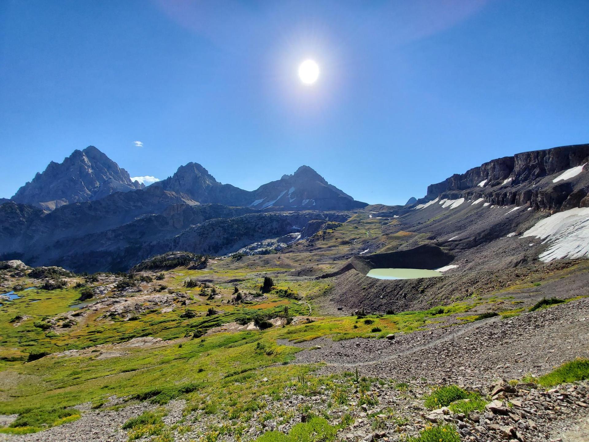 A bright sun shines over a green lake, rocky terrain, and jagged mountain peaks.