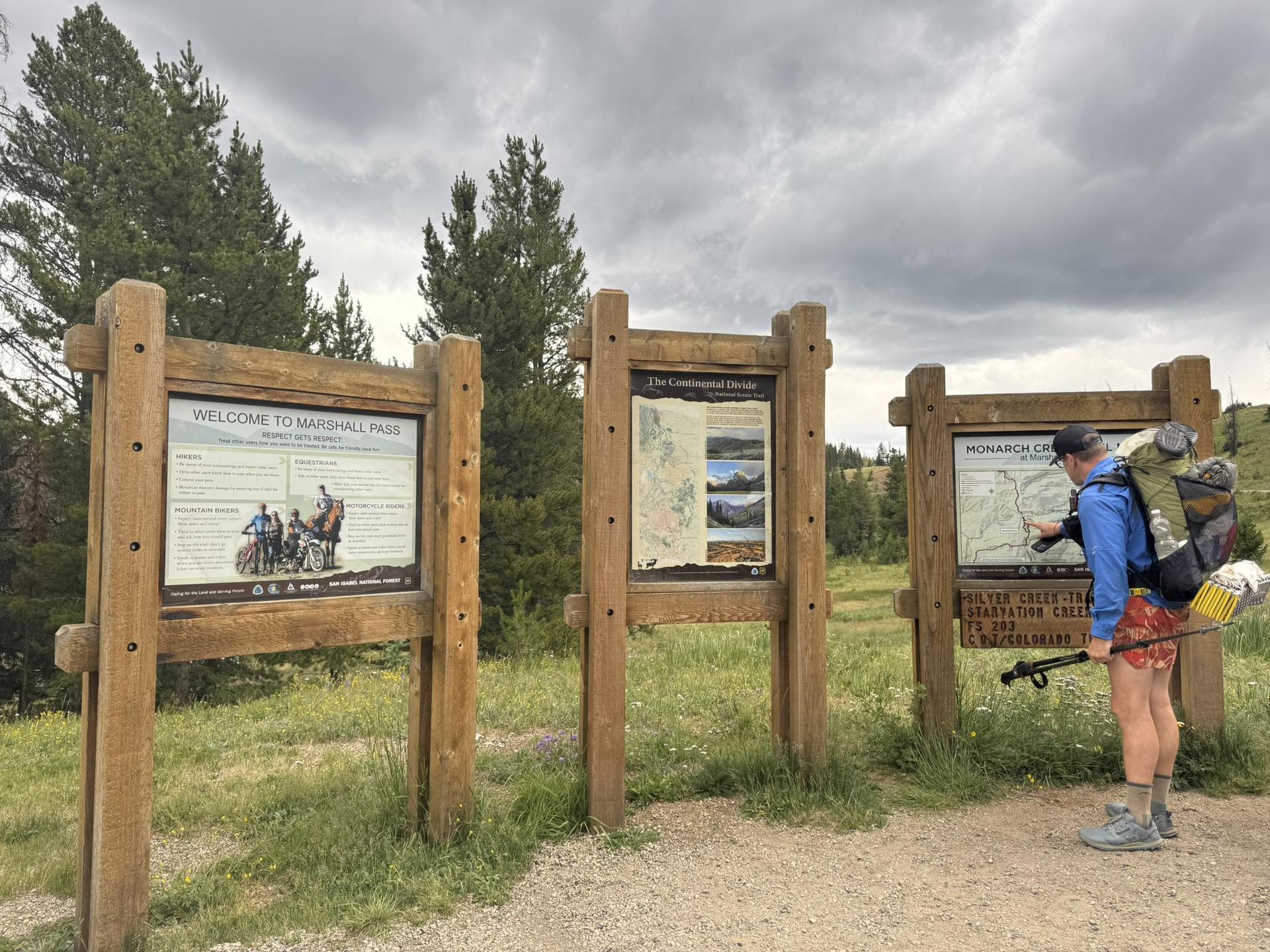 Hiker with backpack reads an outdoor information sign under an overcast sky.