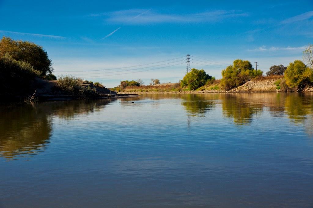 Calm river reflecting bright blue sky, flanked by green trees and power lines.