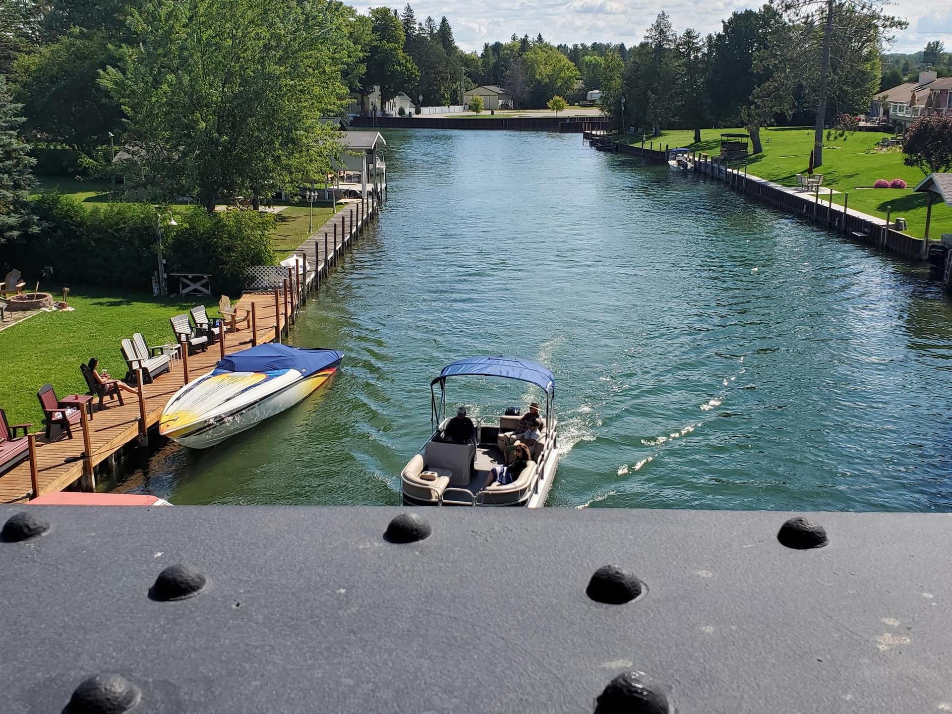A pontoon boat with people moves on a sunny canal past docks and green banks.