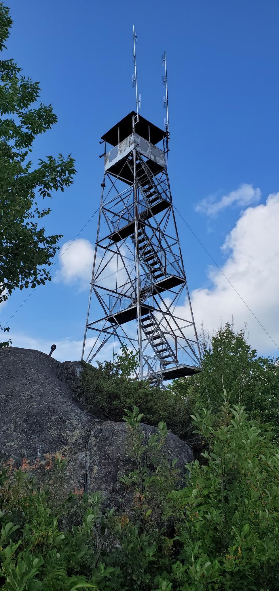 A metal fire lookout tower stands on a rocky outcrop with a person visible.