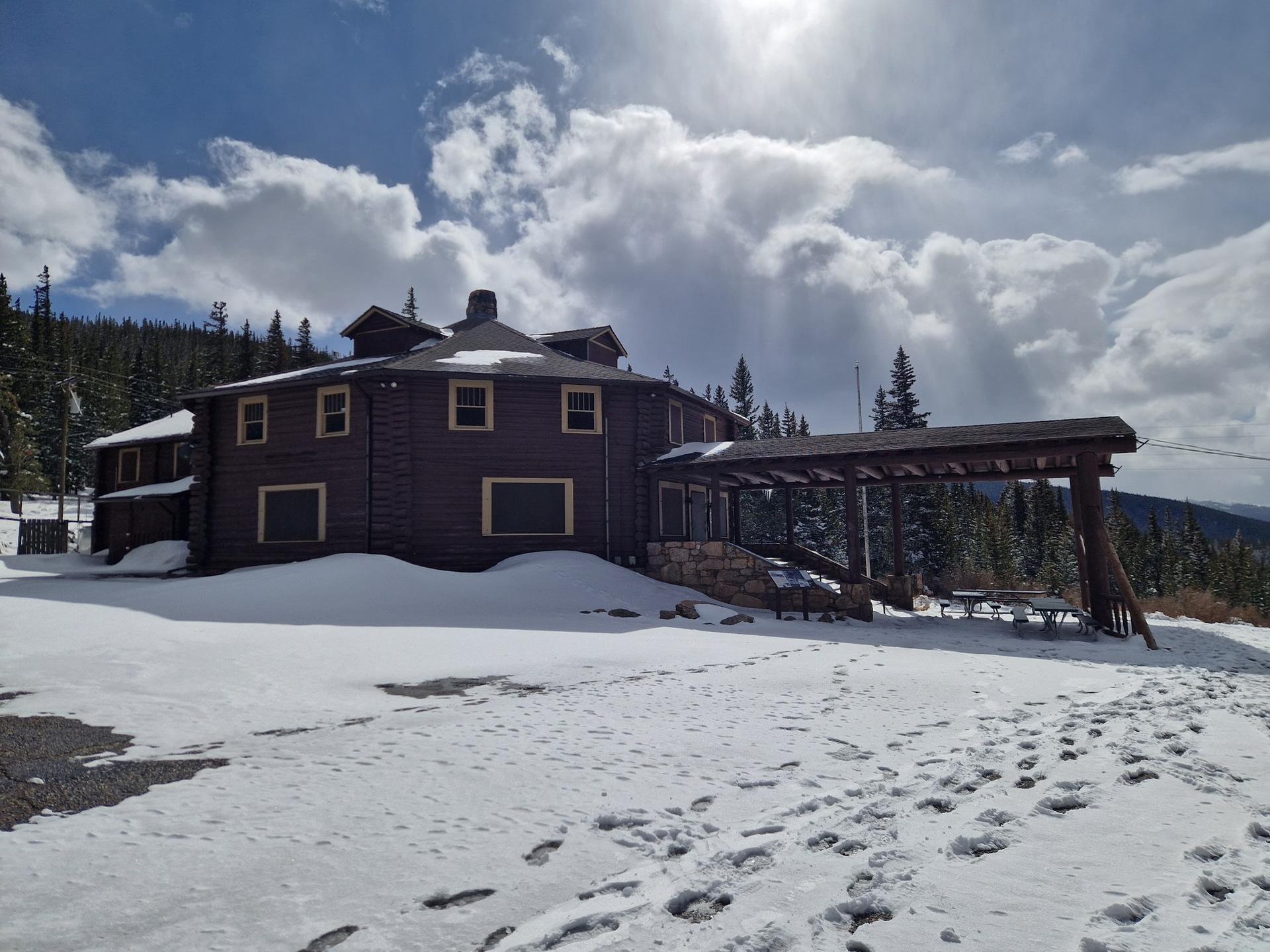 A large wooden lodge stands in a snowy landscape under a bright, cloudy sky.