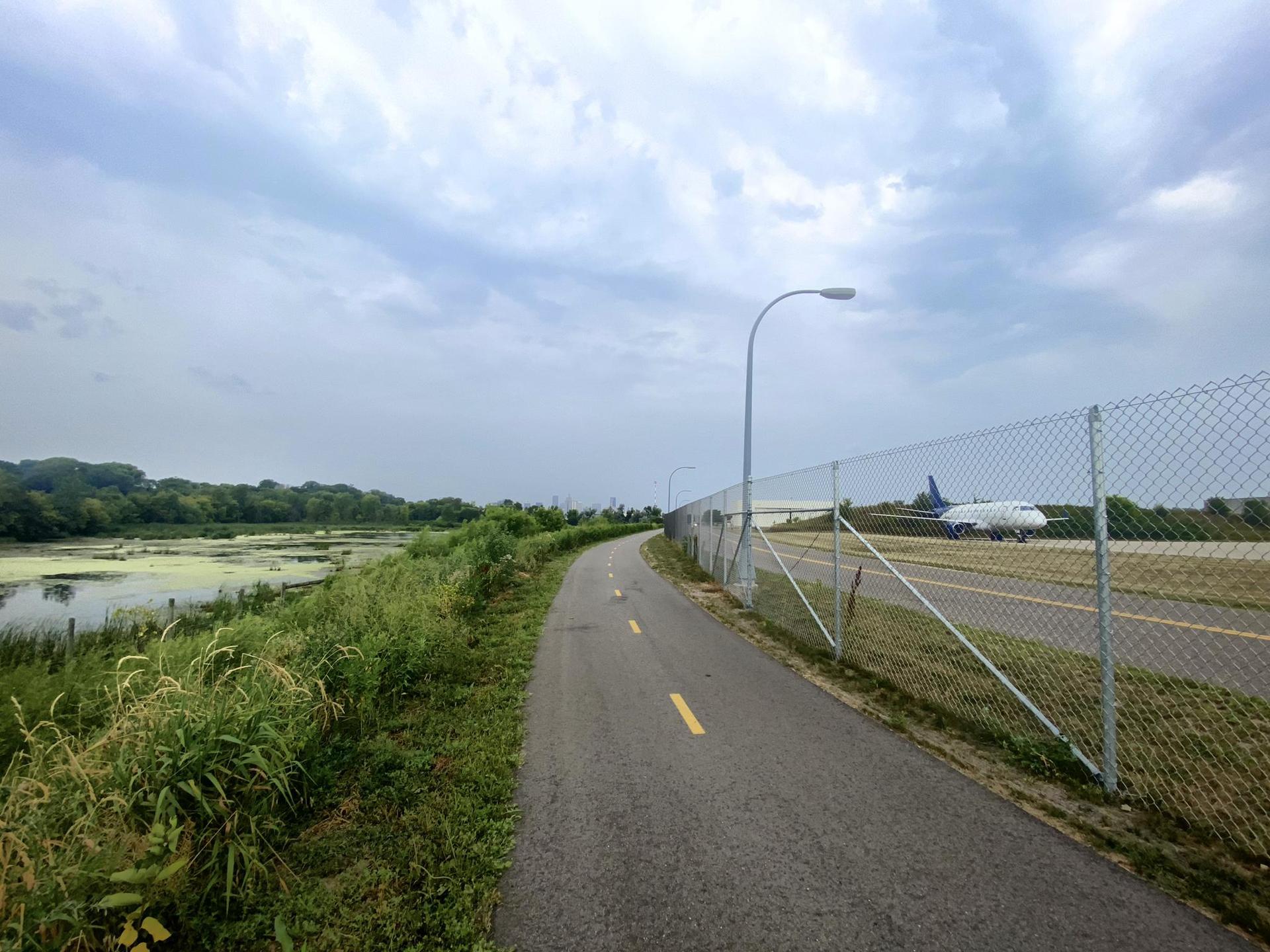 Paved path next to a fence, with an airplane on a runway and marshland.