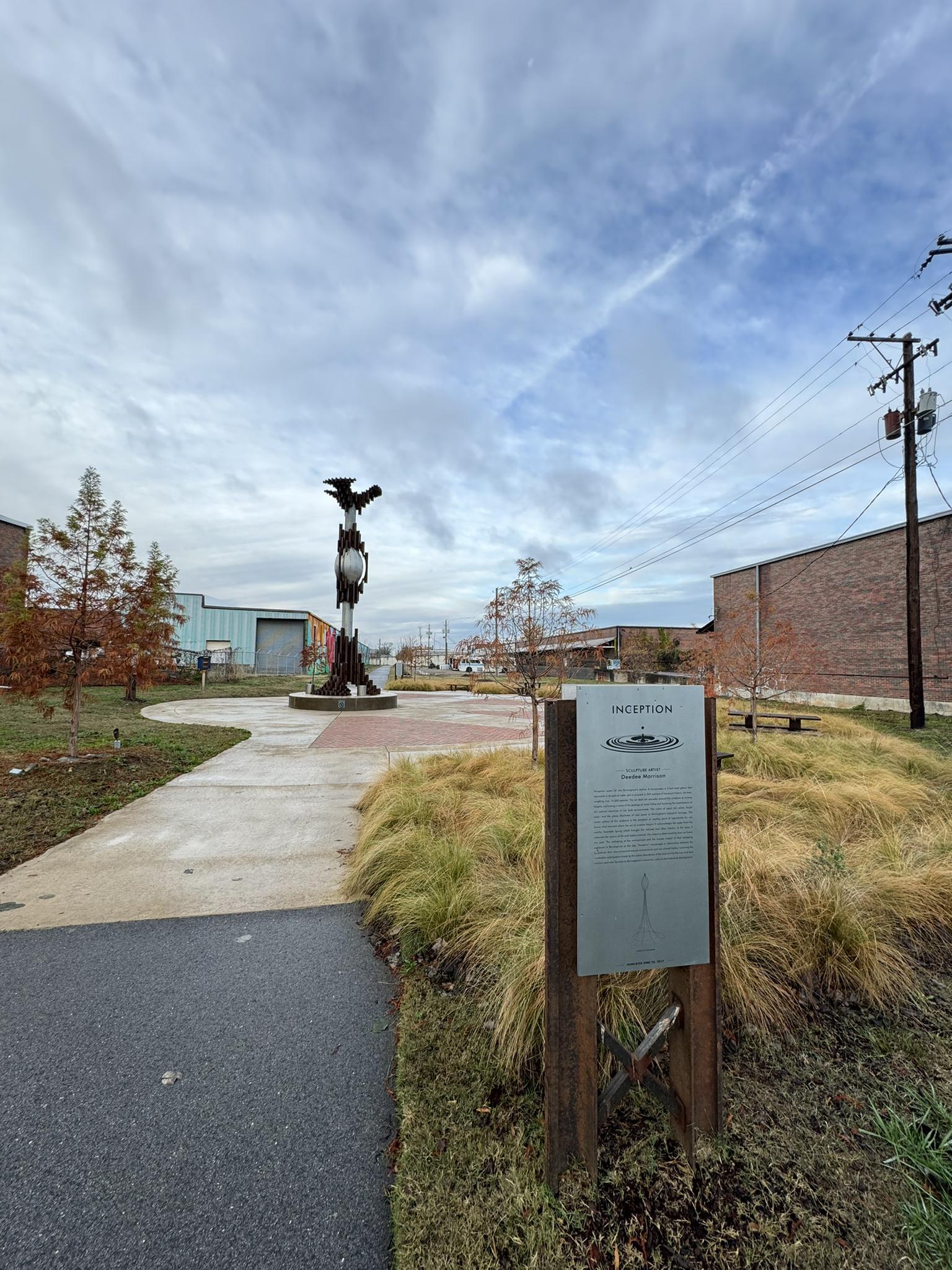Abstract metal sculpture and an informational sign beside a path under a cloudy sky.