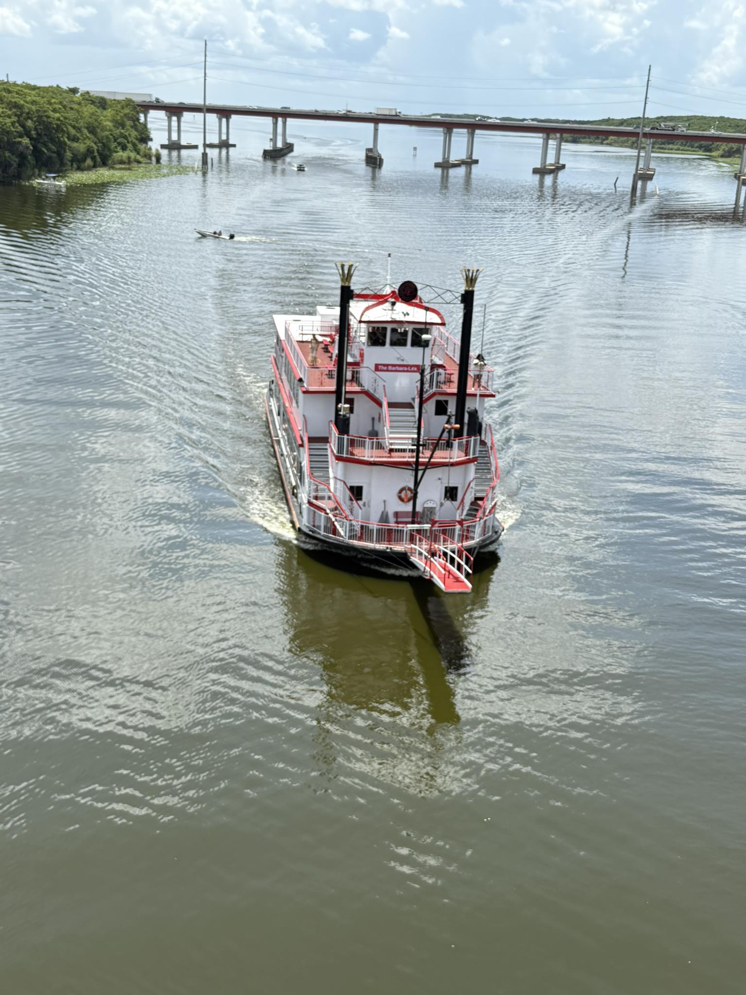 A red and white paddle boat cruises on water under a distant bridge.