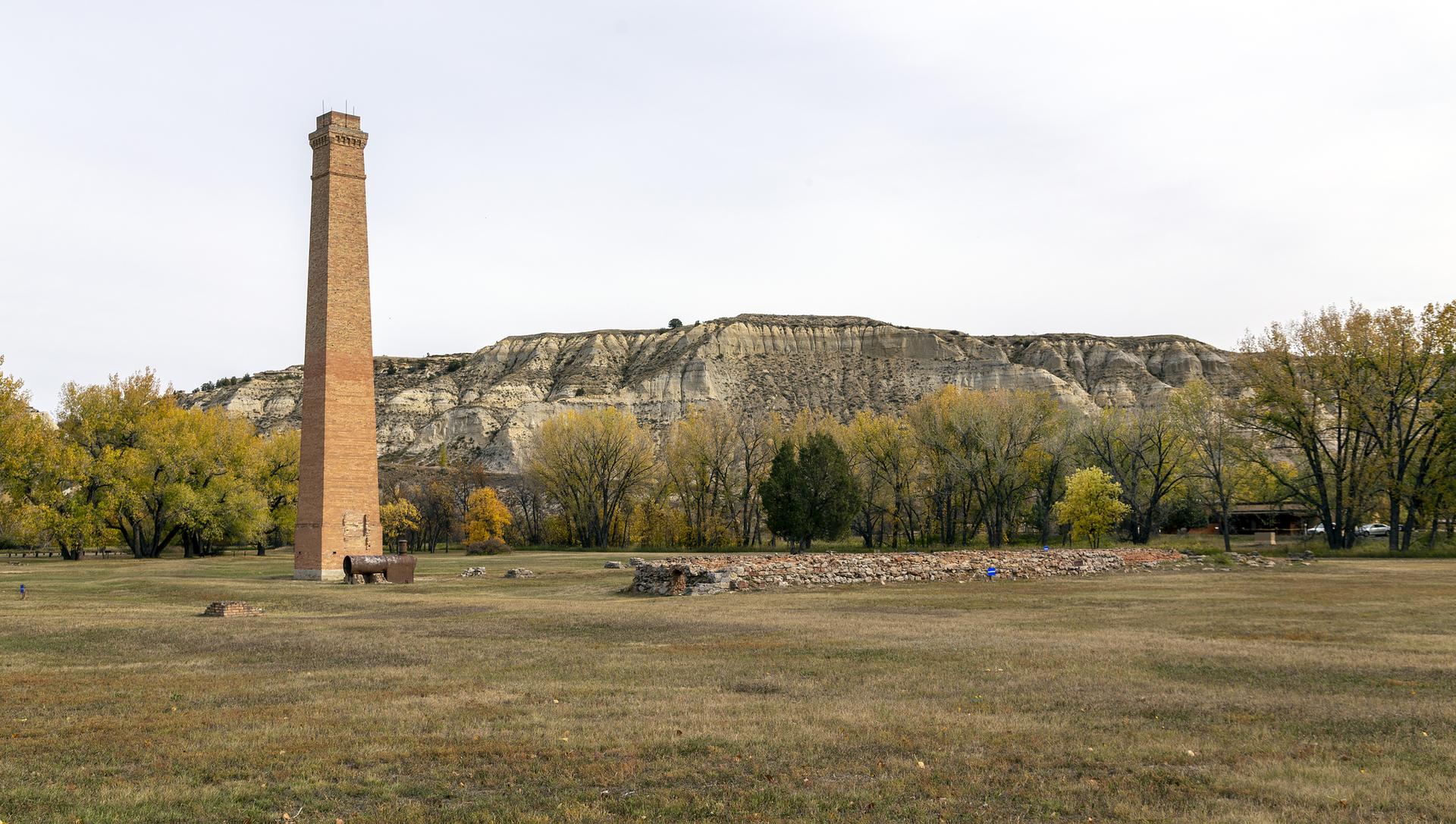 Tall brick chimney in a grassy field with autumn trees and rugged hills.