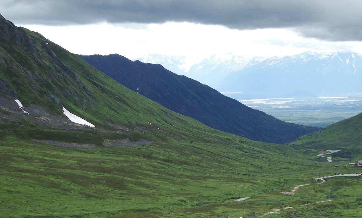 Vast green mountain valley under an overcast sky, with distant snow-capped peaks.