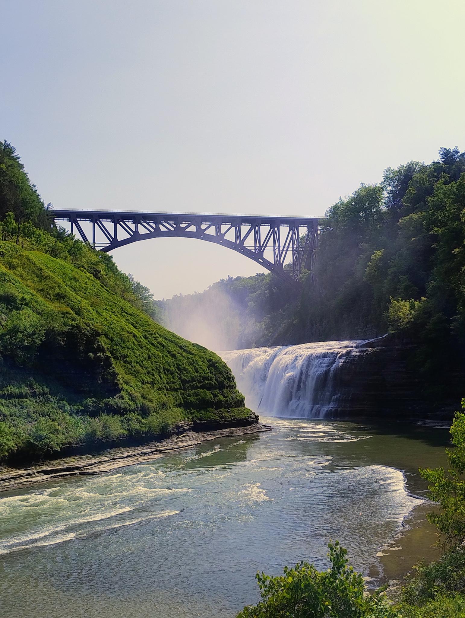 Majestic waterfall and river under a large bridge on a bright day.