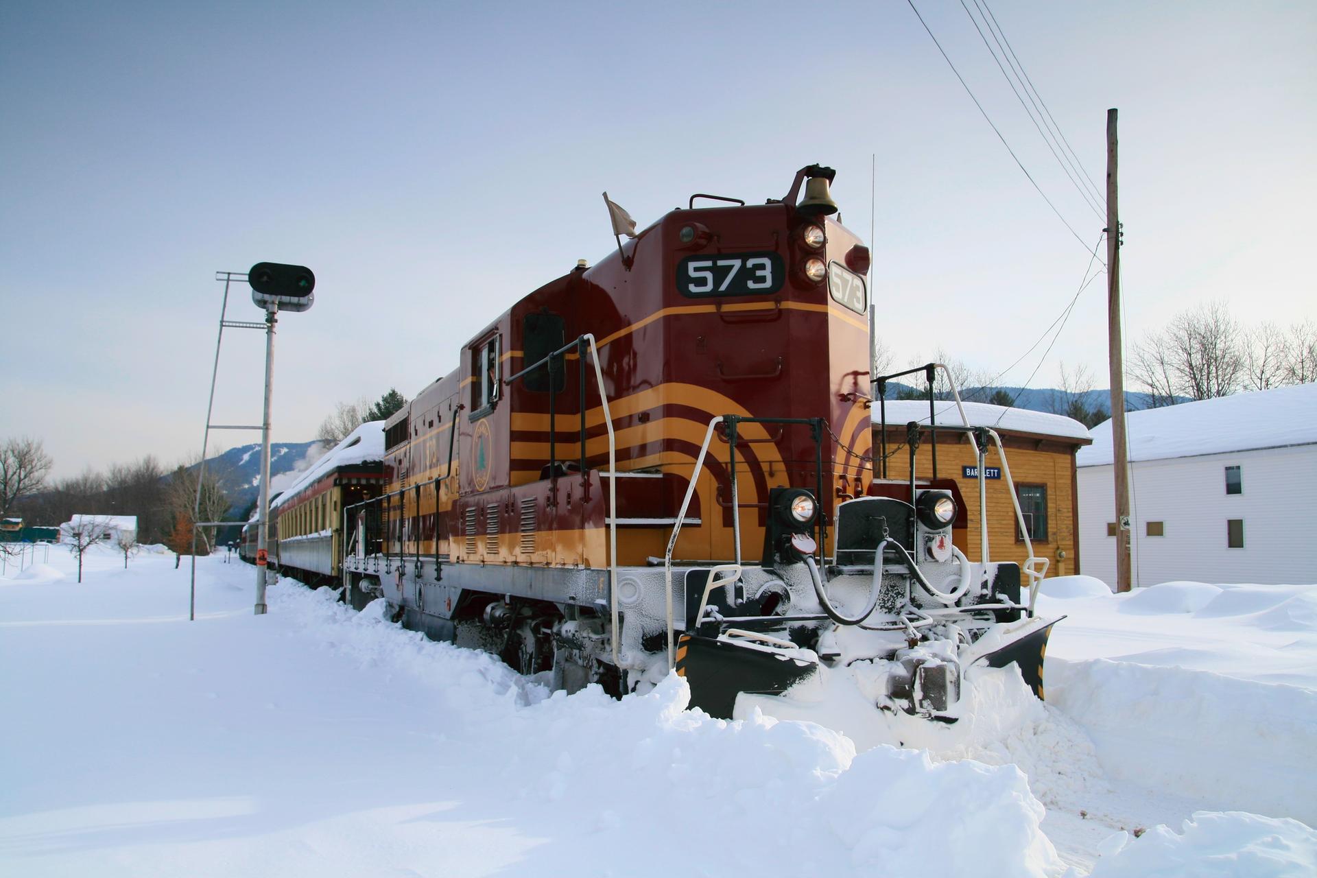 Red and yellow train with snowplow clearing tracks in a snowy winter landscape.