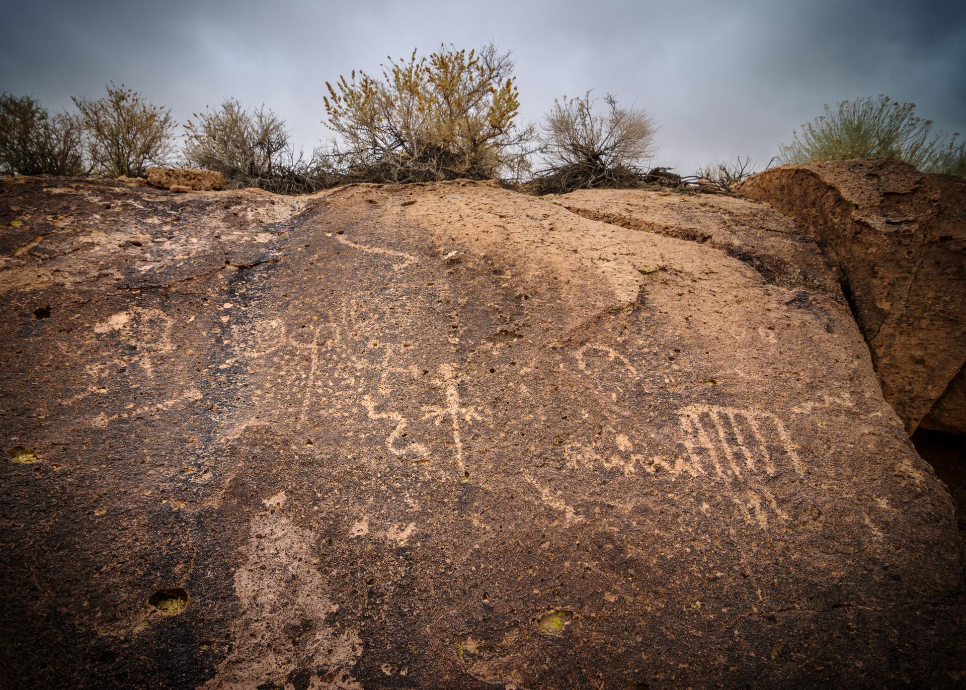 Ancient petroglyphs carved into a large, dark rock under an overcast sky.