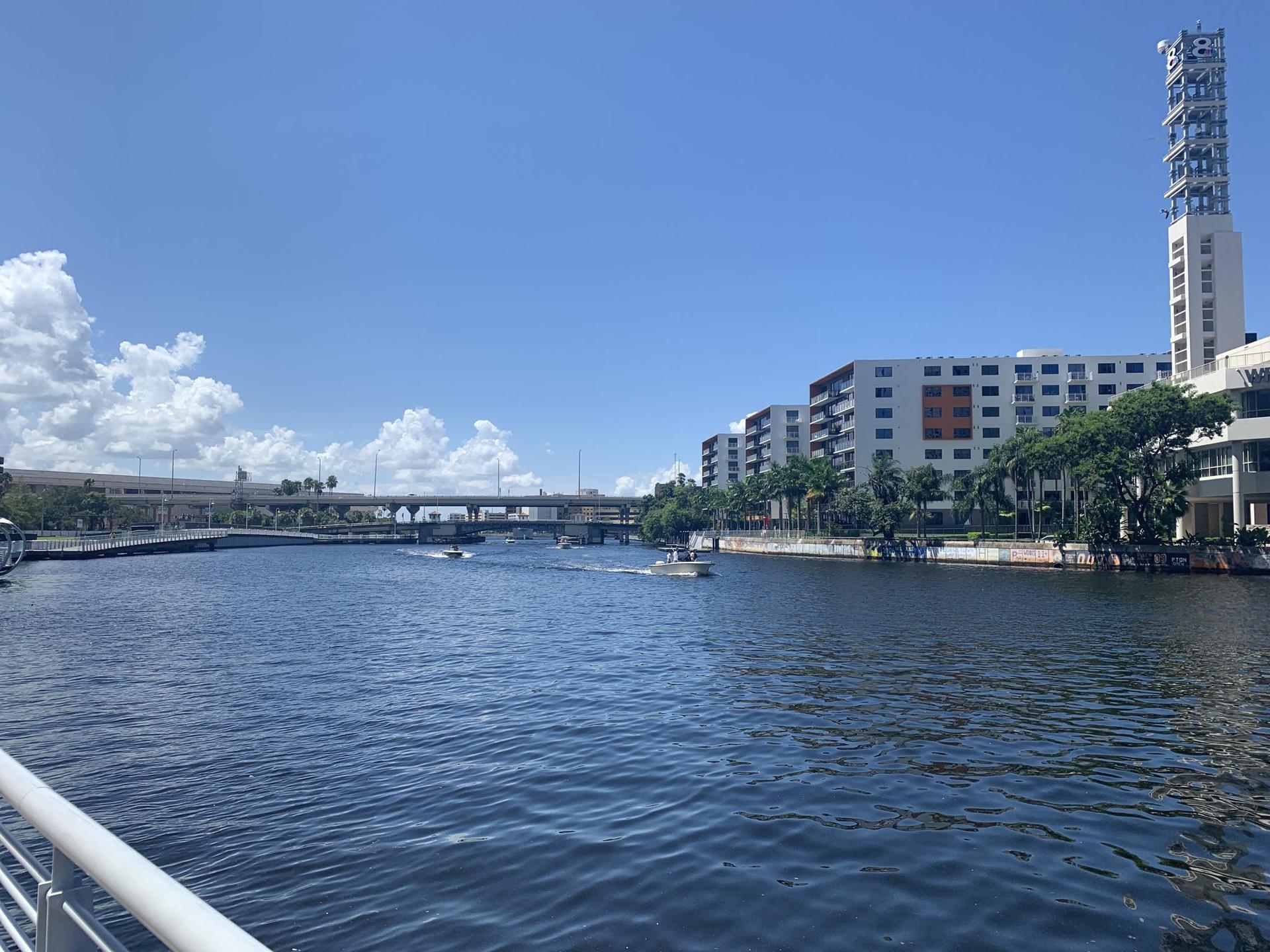 Sunny urban river with boats, a bridge, and tall buildings under blue sky.