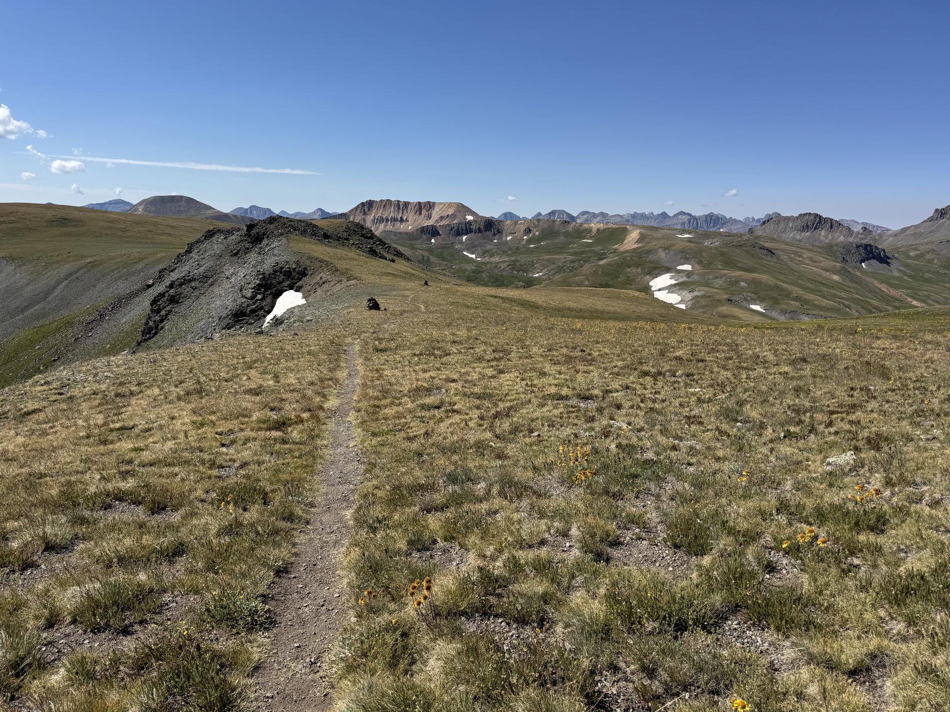 A narrow trail crosses a high grassy ridge with distant mountains and blue sky.