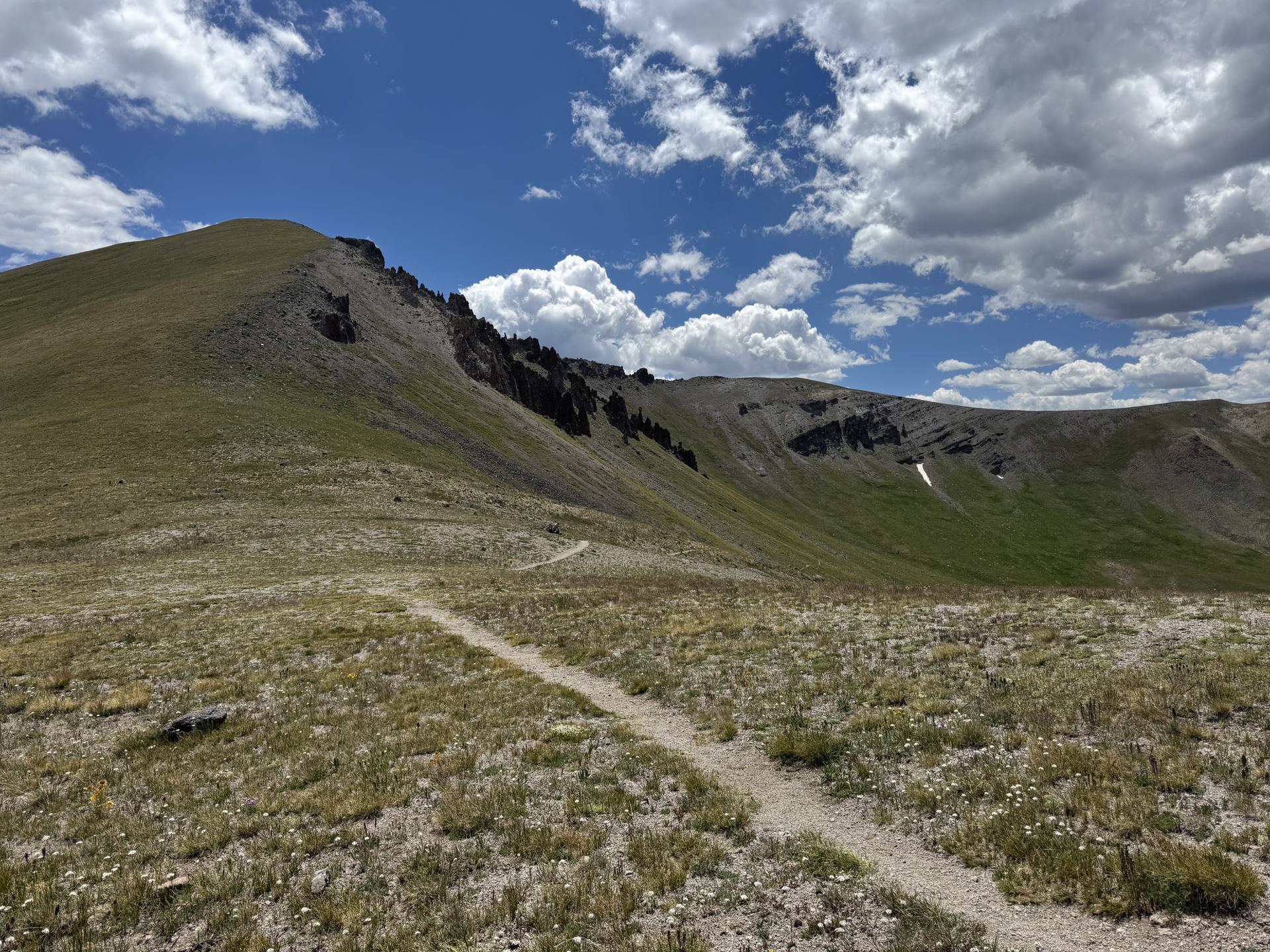 A dirt trail winds up a grassy mountain slope under a bright, cloudy sky.