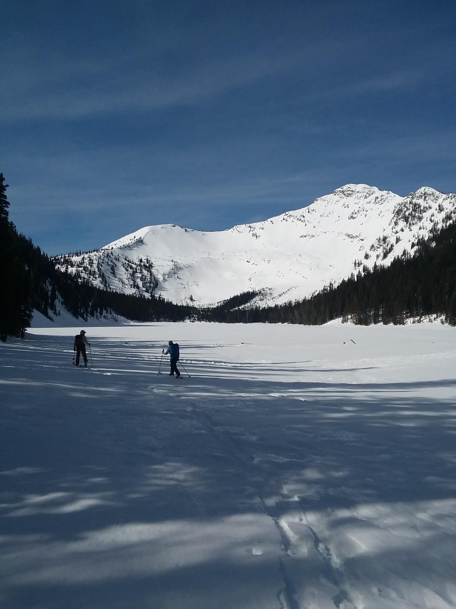 Two people cross-country ski across a sunny, snow-covered valley with mountains.