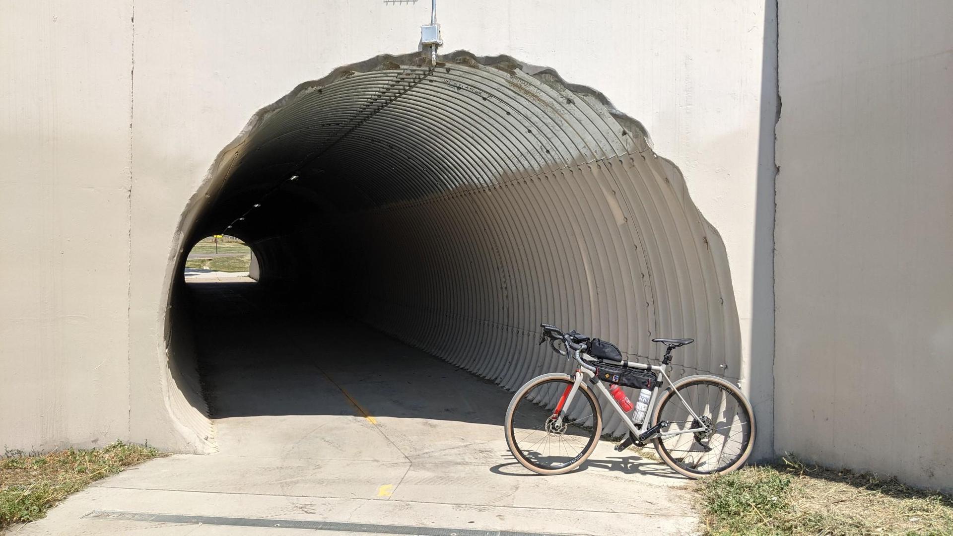 Bicycle parked beside a long, dark corrugated tunnel entrance in daylight.