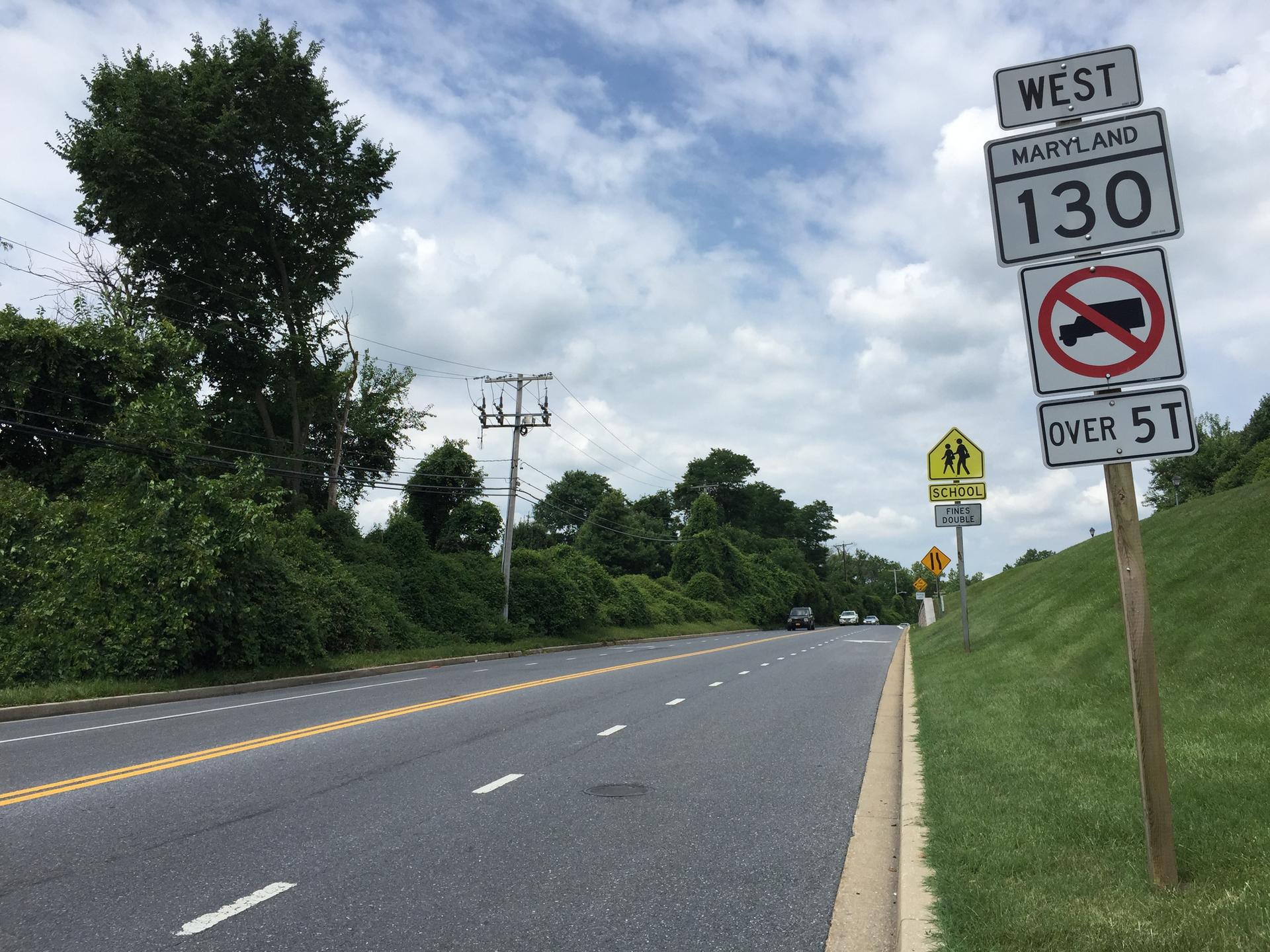 Multiple road signs stand beside a paved road under a cloudy sky.
