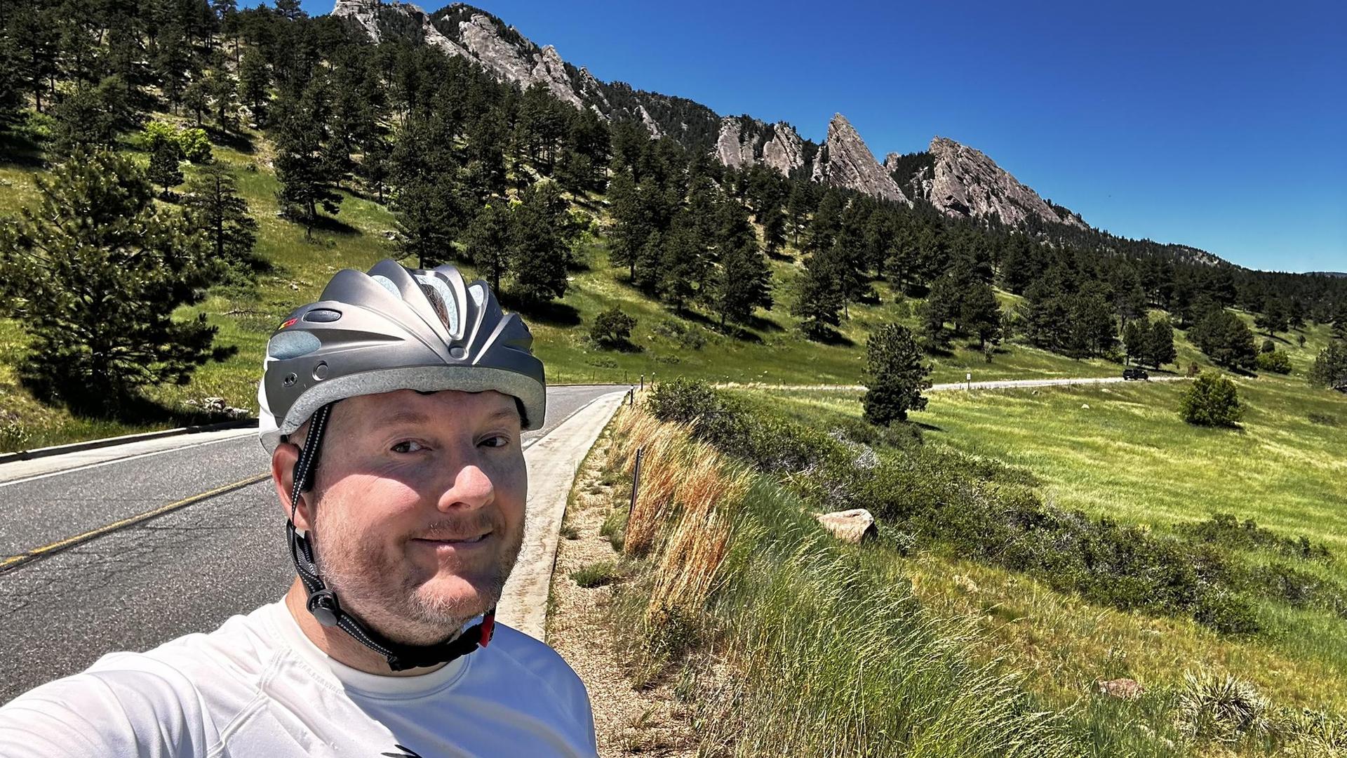 Man in a helmet smiling, with a road and green mountains behind him.