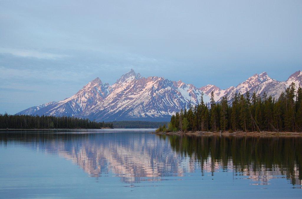 Calm lake reflects snow-capped mountains and a pine forest under soft light.