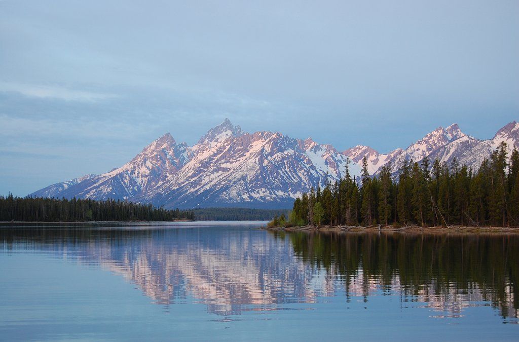 Calm lake reflects snow-capped mountains and a pine forest under soft light. - Great Divide Mountain Bike Route: Two Ocean Lake Loop