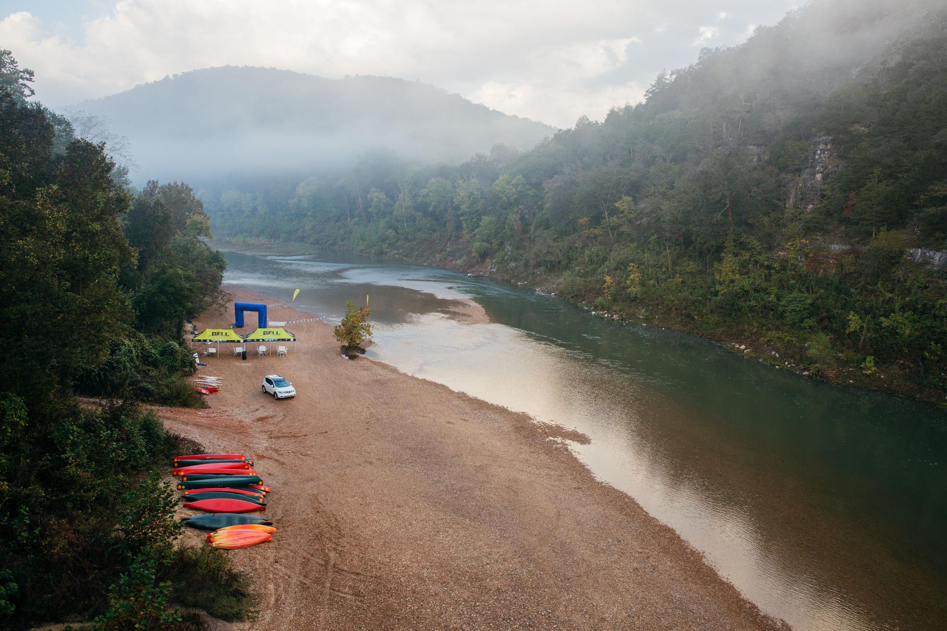 Misty river valley with sandy bank, colorful kayaks, tents, and a car.