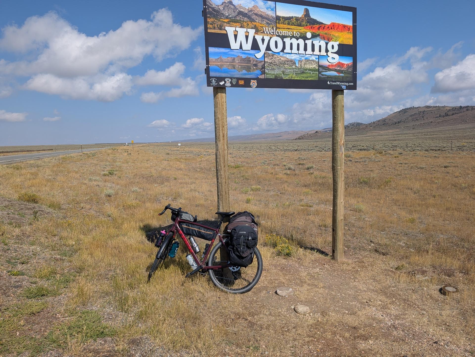 Loaded bicycle parked beside a large welcome sign on a sunny day.