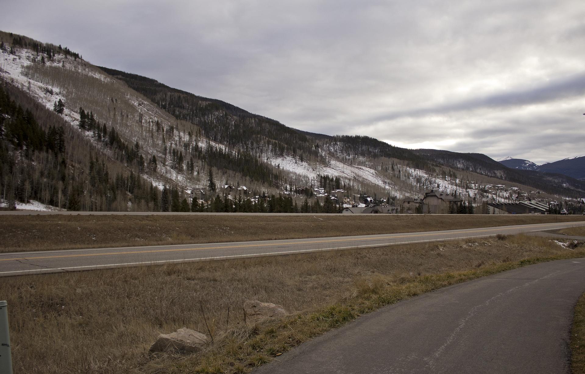 Wide road leading to a snow-dusted mountain with distant buildings under a cloudy sky.