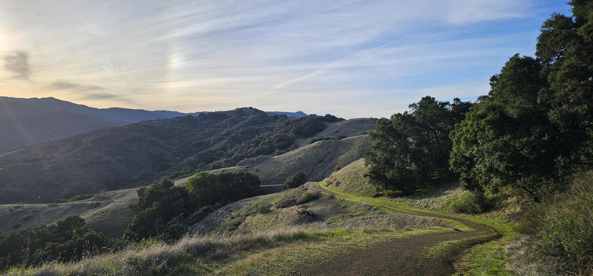 Winding dirt trail on a green hillside with distant mountains at sunset.