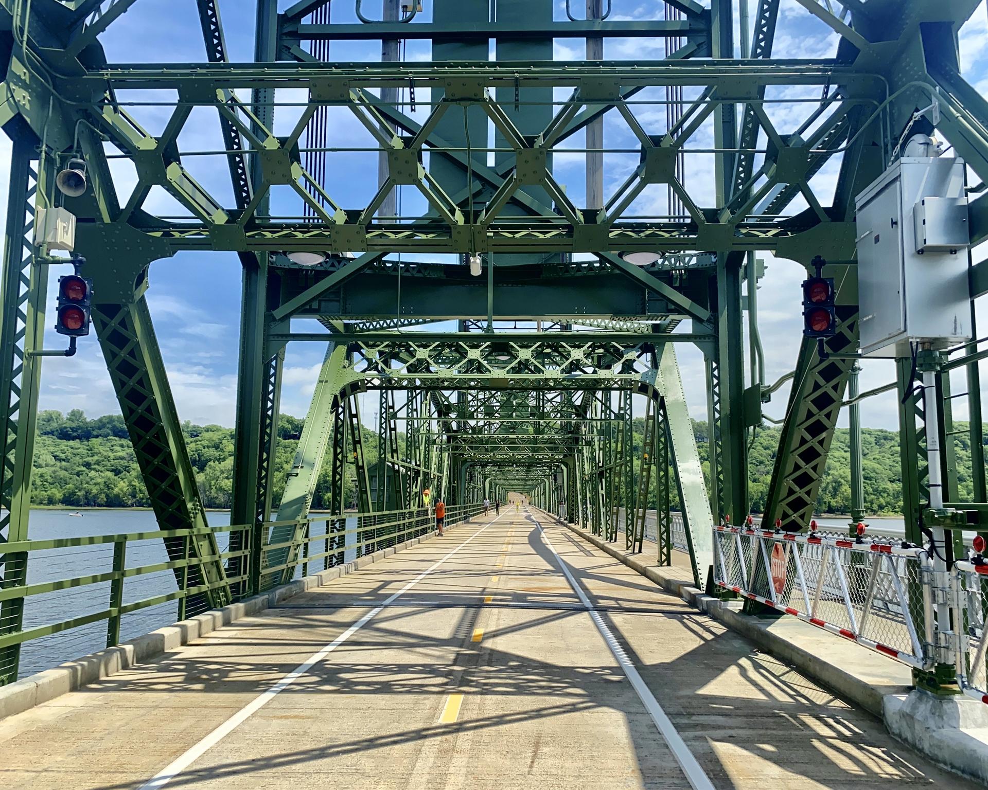 A person walks on a green metal truss bridge over water on a sunny day.