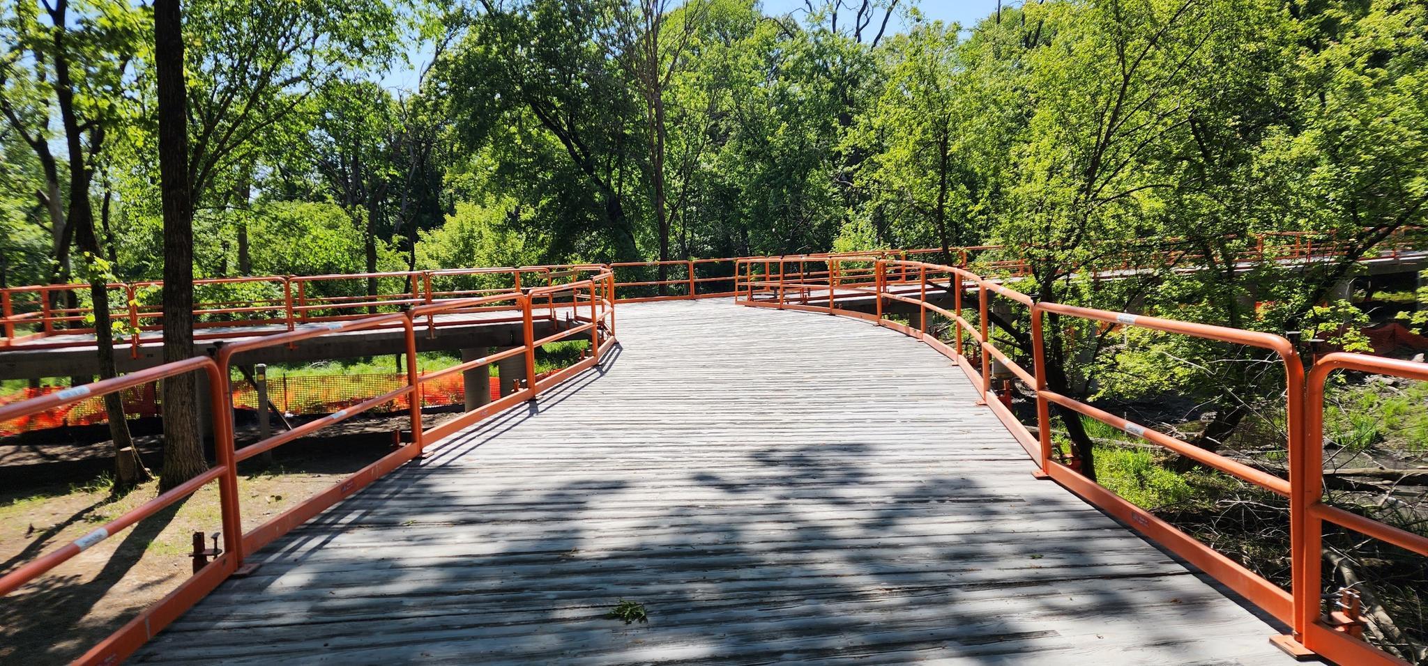 A winding wooden boardwalk with orange railings through a sunny green forest.