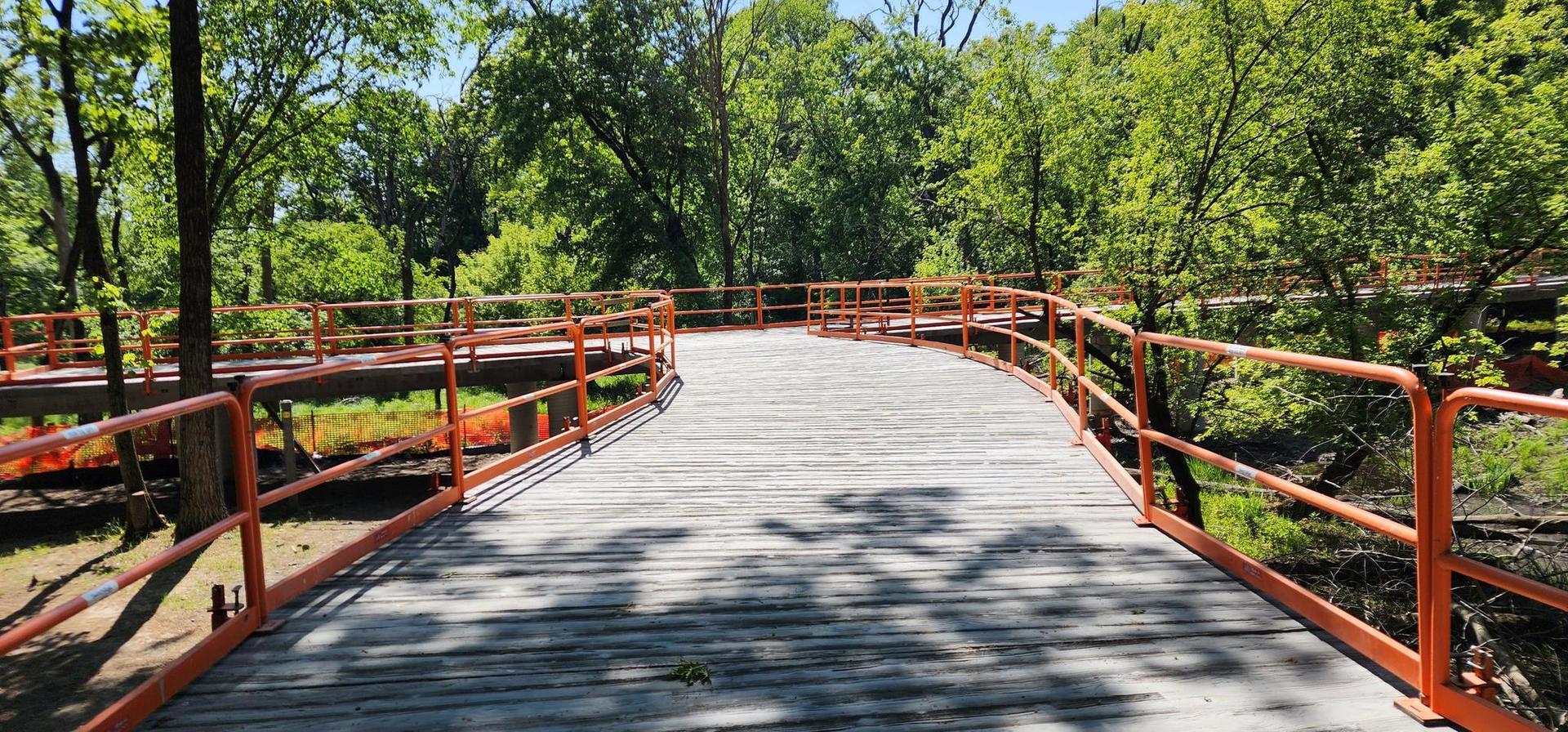 A winding wooden boardwalk with orange railings through a sunny green forest.