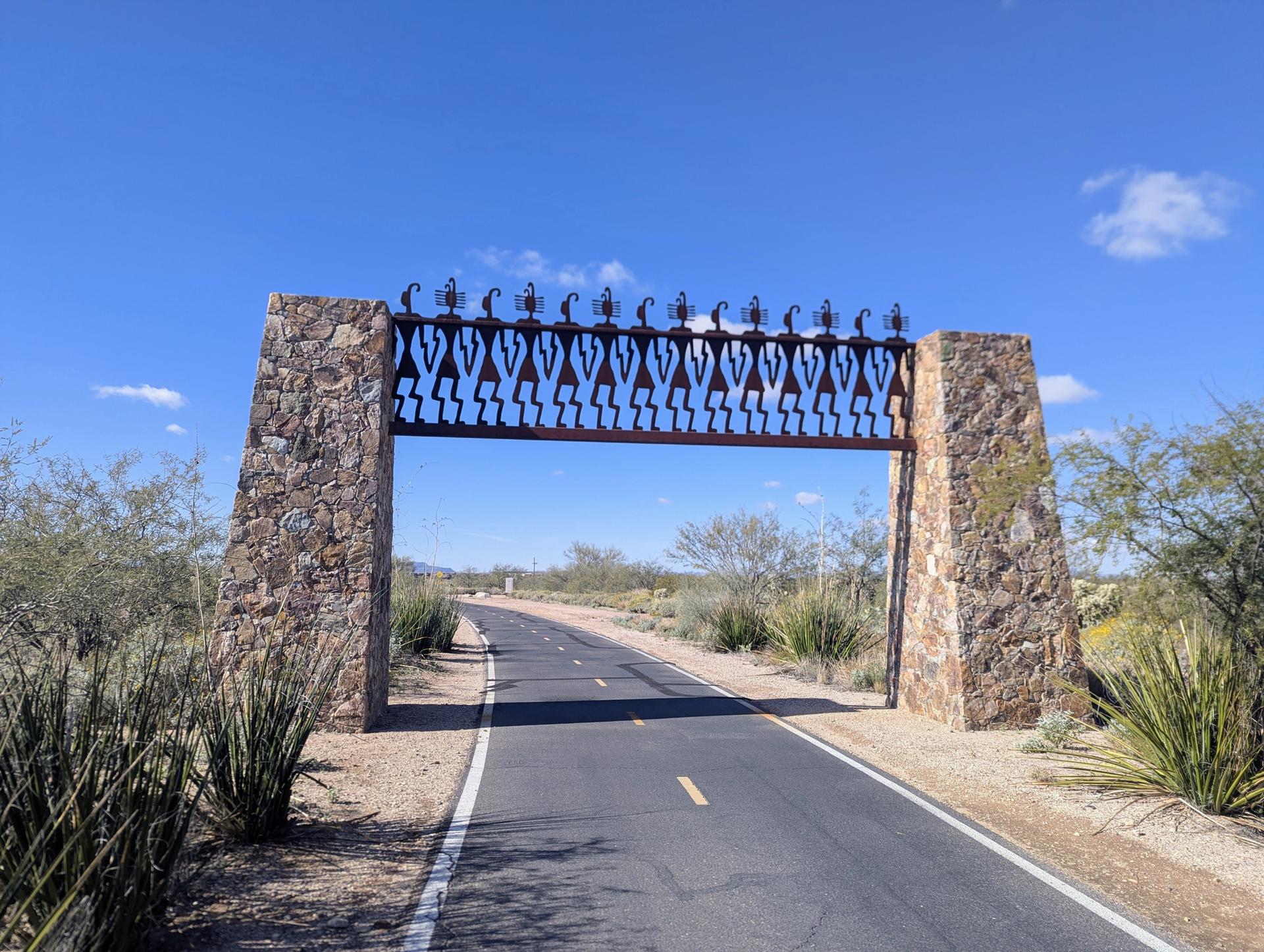 Paved path leading through a decorative stone and metal archway under a clear sky.