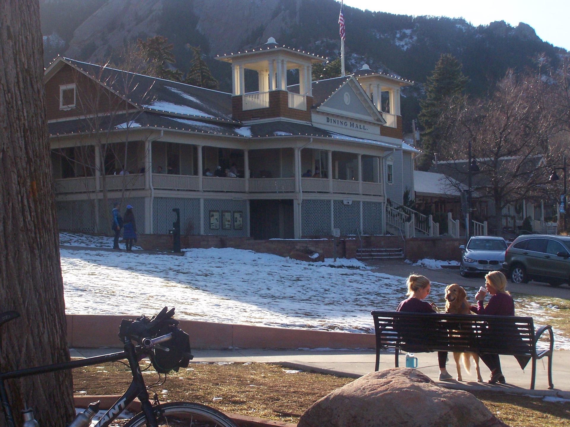 Large building with snow, two women and a dog sitting on a bench.